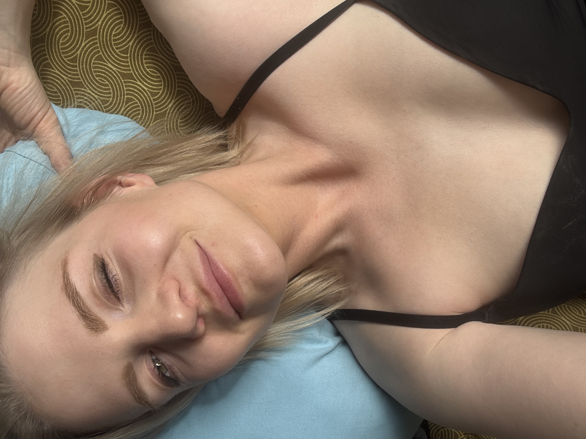 The primary subject of the image is a young woman, captured in a close-up, high-angle shot, lying down in an indoor setting, presumably in Saint Petersburg, Russia. She has blonde hair, light skin, and wears a black strap top or camisole. Her expression is relaxed and content, characterized by a gentle smile and her left eye appearing partly closed, suggesting either a playful wink or a sleepy gaze. Her head is nestled on a light blue pillow or soft fabric, with her left hand visible in the upper left, resting on the blue material. Her collarbones are distinctly visible. The background features a distinctive brown and gold fabric with a repeating pattern of concentric circles, indicating patterned bedding or upholstery. The lighting is soft and even, highlighting her facial features and upper body. No visible text, specific time of day, or weather conditions can be discerned from the image.