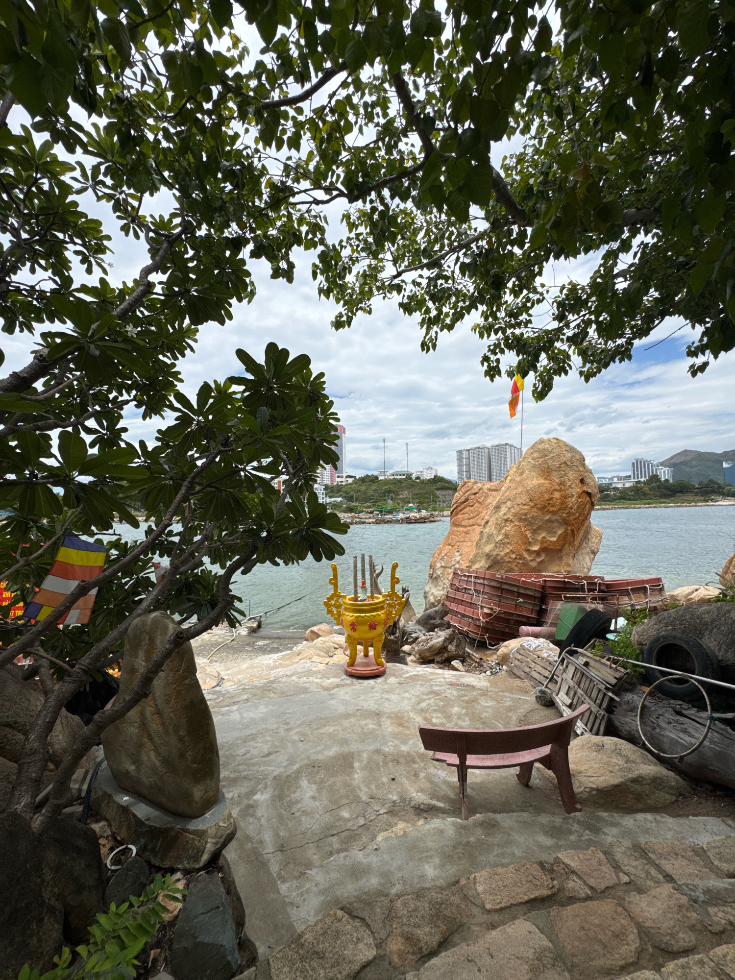 The image depicts an outdoor scene in Nha Trang, Vietnam, likely near the coast, with a body of water visible in the background. The foreground is dominated by lush green foliage from trees arching over the scene, obscuring parts of the sky.
On the ground, there is a concrete patio area. To the left, a small, upright stone object is placed on a larger rock. Further left, a string of Buddhist prayer flags with yellow, red, white, and blue stripes is visible, attached to a tree branch. In the center of the patio, a golden incense burner with intricate designs sits on a red base. Behind the incense burner, a large, rough-textured orange rock formation is prominent.
In front of the large rock, there are stacked items that appear to be red plastic crates or containers, tied together with ropes. To the right of these crates, various objects are scattered, including what appear to be old tires, wooden planks, and other debris. A small, dark red bench with curved legs is positioned on the concrete area in the mid-right of the frame, facing slightly away from the viewer.
The background shows a calm body of water, likely the sea, with a few boats visible in the distance. Beyond the water, a city skyline with several tall buildings and mountains can be seen under a cloudy sky. The overall impression is a somewhat eclectic arrangement of natural elements, religious artifacts, and discarded items in a coastal setting. No people are present in the image.
