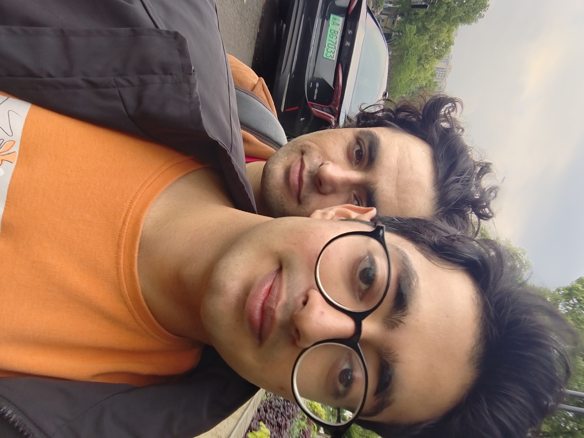 Two men are close together, taking a selfie. The man in the foreground is wearing black-rimmed round glasses and an orange t-shirt, covered partially by a dark jacket. The man behind him has dark curly hair and is wearing a light orange shirt. A black car is partially visible in the background with the license plate 渝 A BG7033. Based on the license plate, the location is Chongqing, China. The surrounding environment suggests a cityscape with trees and buildings in the background. The photo appears to have been taken outside.