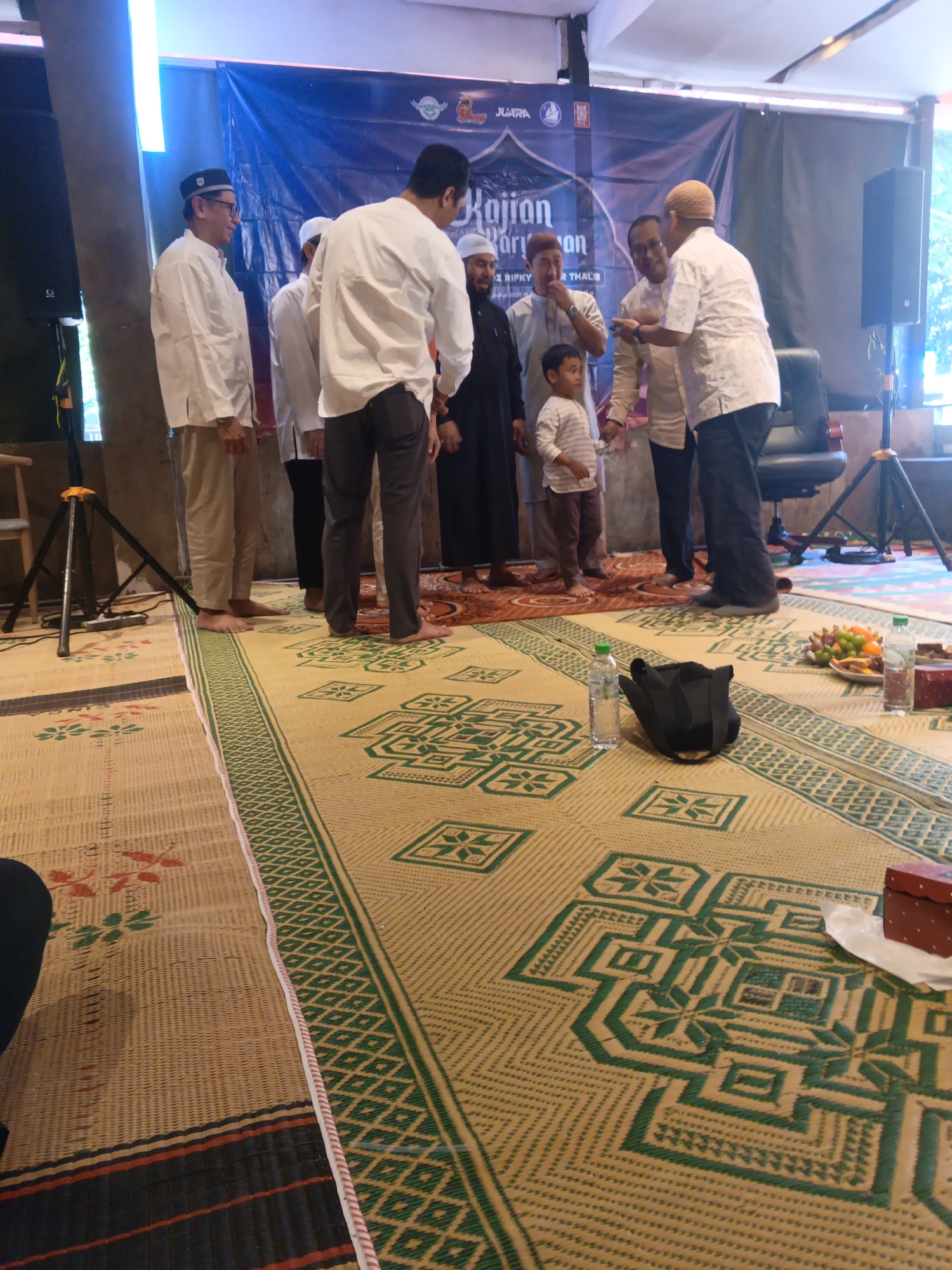 In Surabaya, Indonesia, several men and a young boy are gathered indoors, standing on a large, woven mat. The mat has a patterned design with green and beige tones. Behind them, a blue banner with text and logos is displayed. The men, dressed in traditional Islamic attire including peci hats, appear to be engaged in conversation and possibly an exchange. One man in a white shirt and dark pants is looking towards another man who is offering him something. A black bag and bottles of water are visible on the mat. The environment suggests a religious or community event.