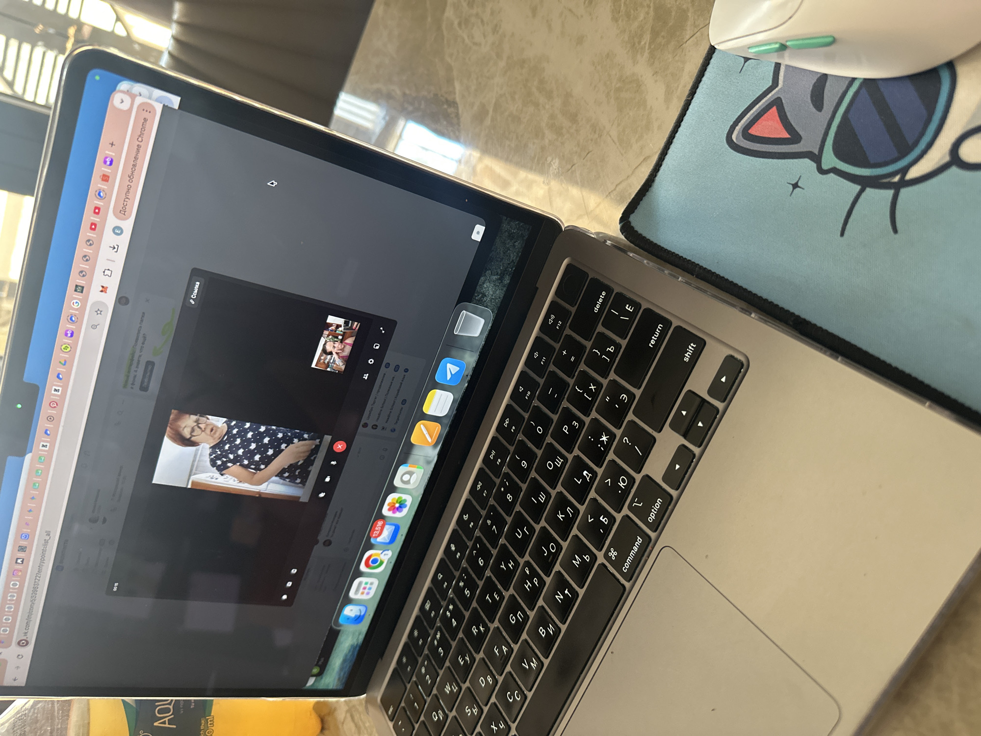 The image shows a laptop open on a table, displaying a video call with a woman visible on the screen. The laptop keyboard has Cyrillic characters. A mouse and a mouse pad with a cartoon cat design are positioned beside the laptop. A bottle of AQUA is partially visible at the bottom left. The presence of Cyrillic on the keyboard suggests the user is potentially in a region with Cyrillic script usage or communicating with someone from that region. The exact location in Bac Nha Trang Ward, Vietnam is not discernible from the image.