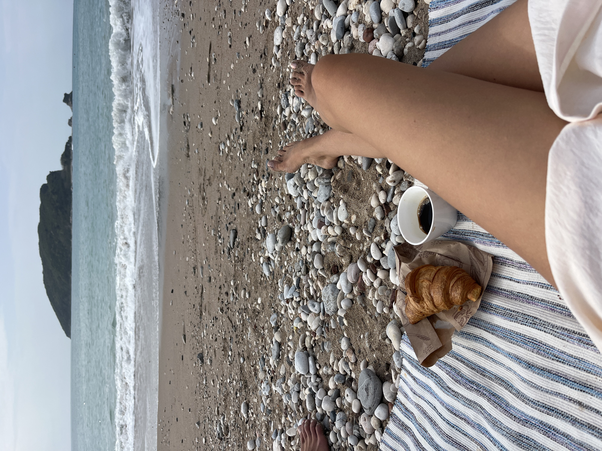 The image shows a woman relaxing on a beach, with her legs crossed. A cup of coffee and a croissant in a paper bag are next to her on a blue and white striped blanket. The beach is pebbly, with the sea and a rocky headland visible in the background. The location is likely near Budva, Montenegro, given the typical Montenegrin coastline characteristics.