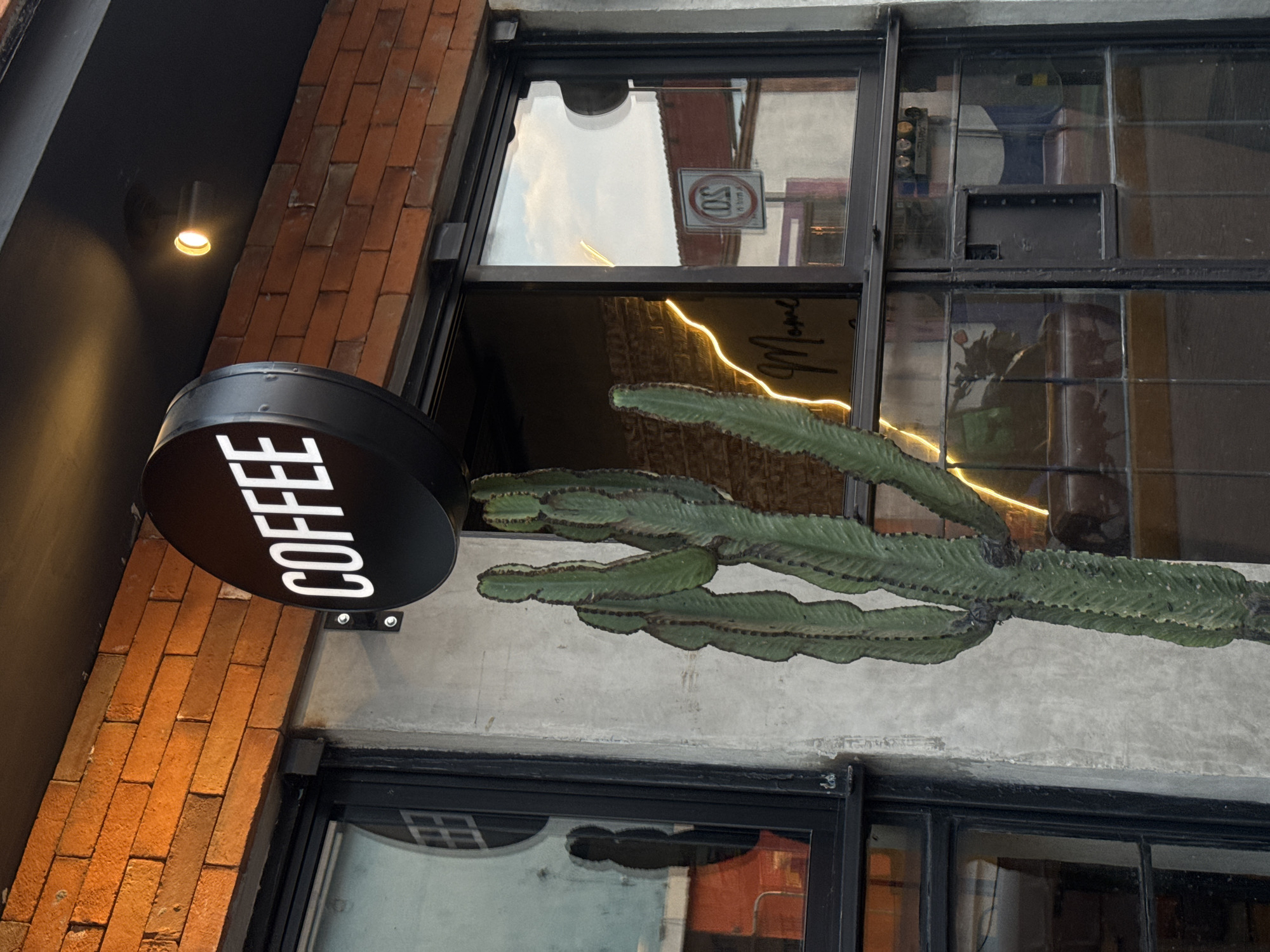 The image captures the exterior facade of a coffee shop, taken from a slightly upward and tilted angle. A prominent round, black sign with the word "COFFEE" in white capital letters hangs from the building, positioned against a section of reddish-brown brickwork on the left and a dark, possibly metallic, overhang above.
A large, green succulent plant, resembling a multi-branched euphorbia or cactus, is affixed to a textured, light grey concrete wall section that forms part of the building's facade. This plant extends horizontally across the frame. Above the concrete and succulent are large glass windows with dark frames, reflecting a pale, possibly overcast, sky and elements of the surrounding environment.
Through the windows, a warm-toned neon light strip can be seen inside, forming a partial word that appears to be "Momo" or "Mom's." Another interior detail visible is a black speaker or vent system integrated into the window frame. Reflected prominently in one of the upper window panes is a circular regulatory sign, featuring a red circle with a diagonal line, containing the number "30" and the word "SOLO" or similar text below it, likely indicating a "No stopping" or "No parking" restriction.
Above the "COFFEE" sign, a single warm-glowing downlight illuminates the dark ceiling area. The mix of brick, concrete, and large glass windows suggests a modern, urban aesthetic for the coffee shop, located in San Andrés Cholula, Mexico. The presence of internal lights suggests the photo was taken during the late afternoon or early evening.
