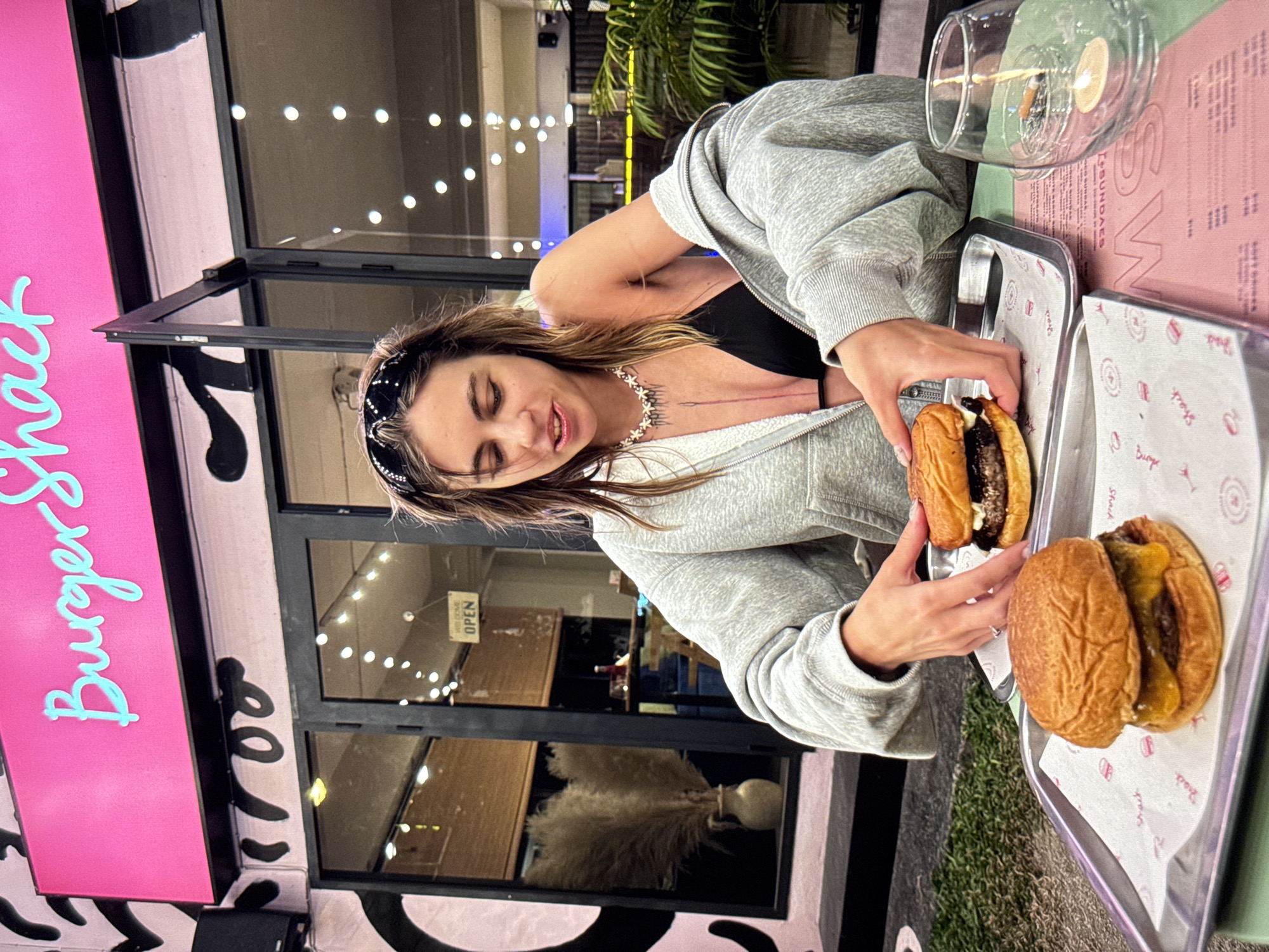 A young woman with long, brown hair and a black bandana is sitting at an outdoor table in front of a restaurant with a bright pink sign that reads "Burger Shack." She is wearing a grey zip-up hoodie over a black bikini top and a star-shaped necklace. The woman is holding a burger in her hands, which is placed on a metal tray with a white paper liner. Another burger is on a similar tray in front of her, and a clear glass with a lit candle and a cigarette butt is to the right. The restaurant appears to have large glass doors and windows, with decorative lighting visible inside. A tall, dried pampas grass arrangement in a vase is seen through the window. The overall setting suggests an evening meal at a casual dining establishment. The location of the Burger Shack restaurant is not specified in the image.