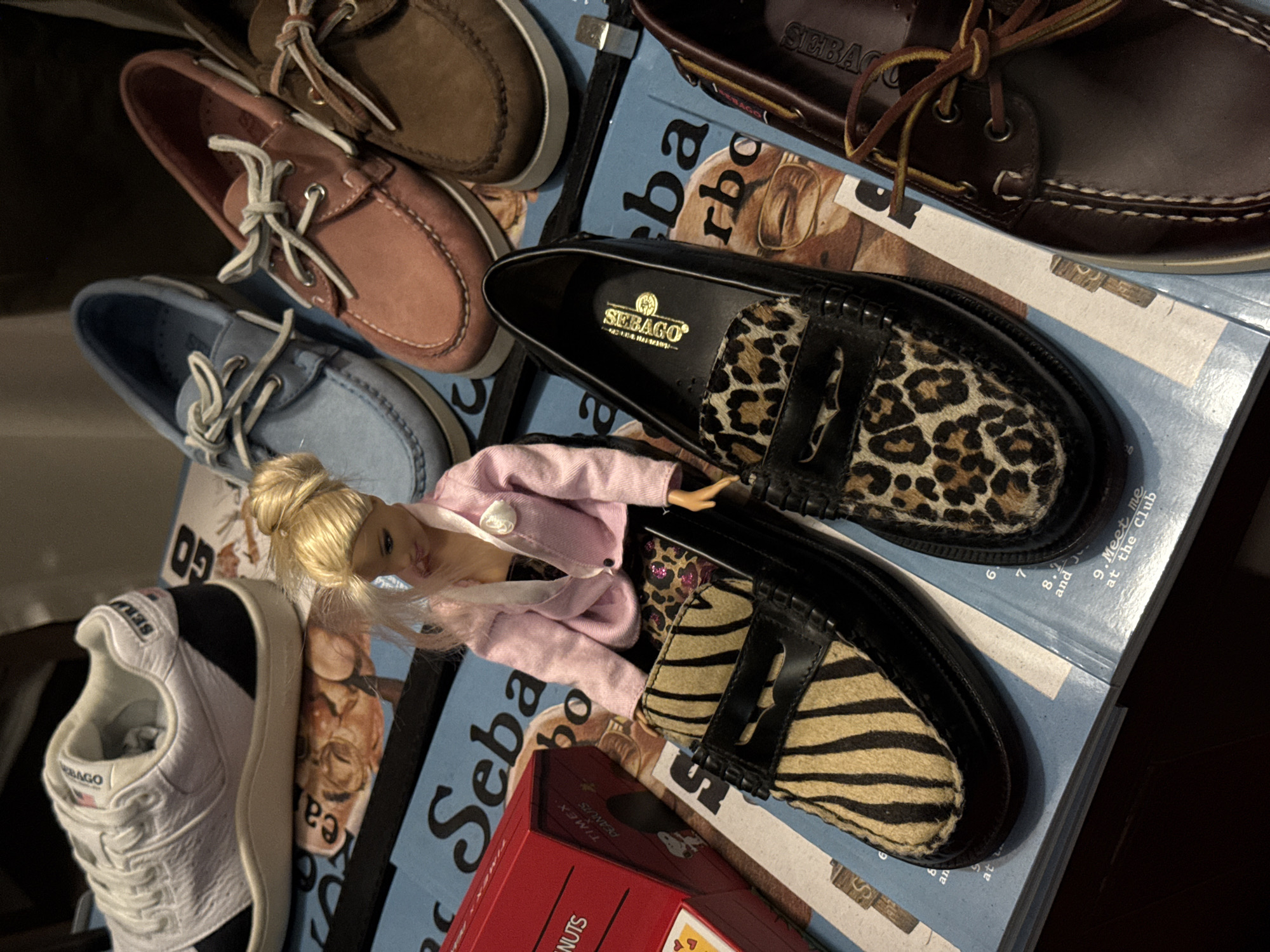 A high-angle close-up shows an assortment of Sebago brand shoes, stacked on what appears to be a stack of Sebago catalogs. There are boat shoes in brown, pink, and blue. A pair of loafers with animal print details and black accents lie atop the catalog, with a blonde Barbie doll posed in between the shoes. A red box, possibly a product container, is also present. The catalogs have large Sebago brand name prints and images. A Sebago sneaker is also present. The scene's environment is dark and appears to be indoors. The city of Milan, Italy, is not represented.