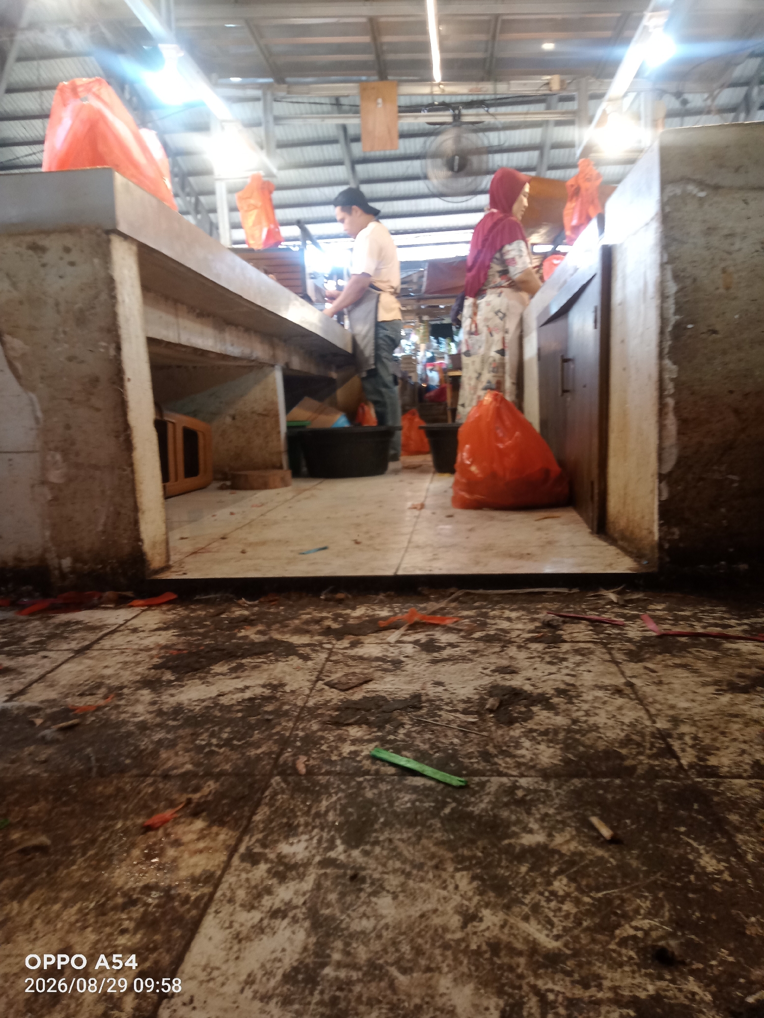 The image depicts a scene inside a market in Jambi, Indonesia. The foreground is dominated by a dirty, tiled floor with scattered debris, including orange and red plastic pieces, and what appear to be dried organic remnants. In the mid-ground, two individuals are visible. A person wearing an apron is standing behind a counter, appearing to be working. To their right, a person in a red headscarf and a patterned dress is also behind a counter. Large orange plastic bags are present on the counter and on the floor. The background reveals a ceiling with corrugated metal and fluorescent lighting. A ceiling fan is visible on the left side of the image. The overall environment appears to be a functional, albeit somewhat untidy, market stall.