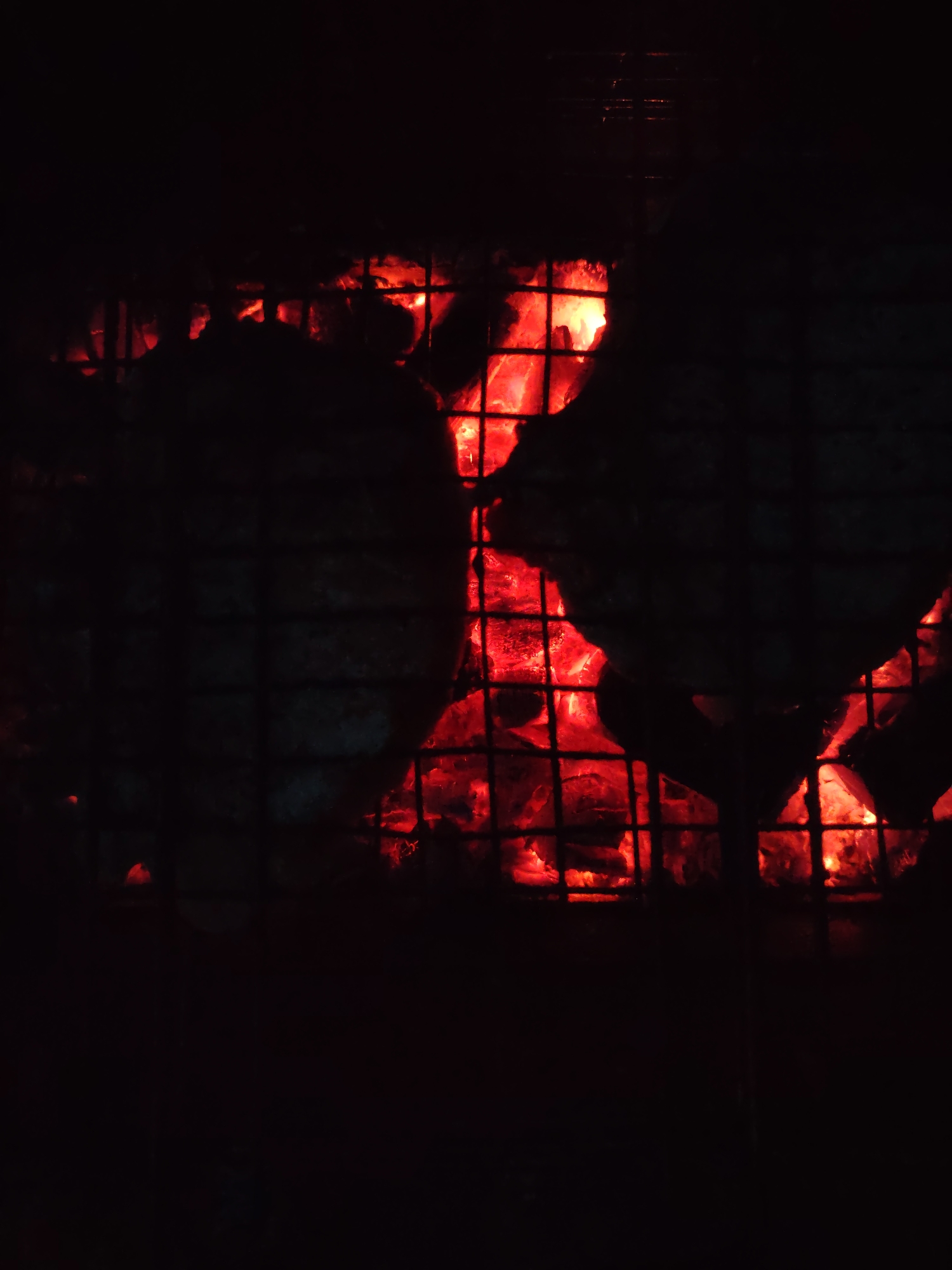 The image depicts a close-up, low-angle shot of glowing red embers and charcoal behind a dark metal grill. The embers are intensely bright, illuminating the space between the grill grates with a fiery orange and red light. The grill itself is a dark, metallic grid pattern that partially obscures the view of the embers. The background is almost entirely black, creating a strong contrast with the glowing coals. There are no people visible in the image, nor any clear indication of a specific location beyond the visual of a barbecue or fire pit. The scene is static and appears to be a detail shot of a heat source.