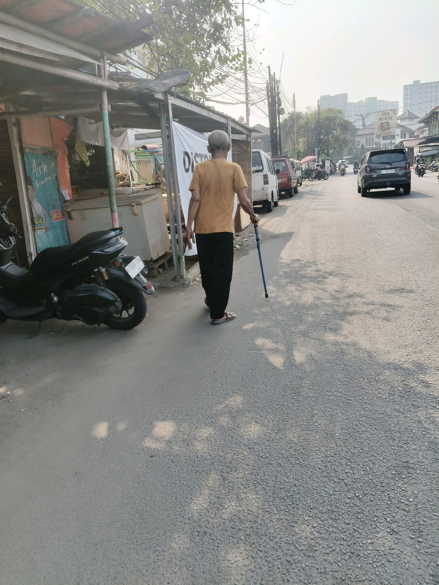 An elderly man wearing a yellow t-shirt and dark pants walks along a paved road in South Tangerang, Indonesia. He is using a blue cane for support. To his left, there is a small roadside stall with a black motorcycle parked in front of it. The stall has a corrugated metal roof and various items displayed. The road is lined with buildings and trees. In the background, several cars and motorcycles are visible on the road, and tall apartment buildings are in the distance. The sky is bright, suggesting it is daytime.