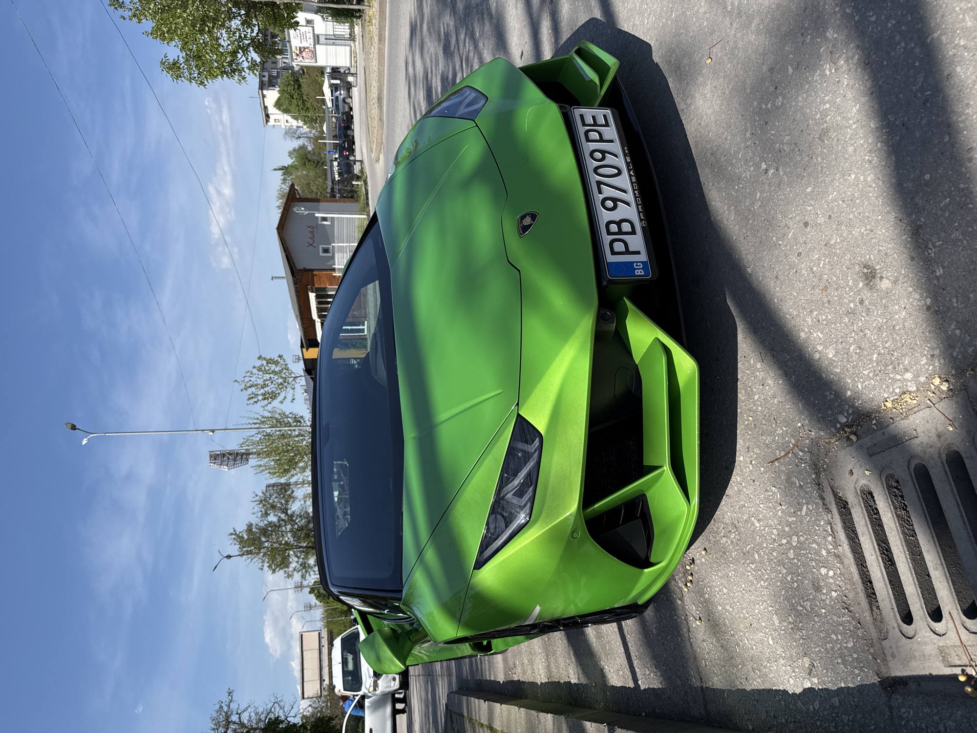 The image displays a vibrant, bright green Lamborghini sports car, likely a Huracán, parked on the right side of an asphalt road under clear, sunny skies. The car is angled slightly, presenting its aggressive front end and a portion of the hood, with its striking color illuminated by direct sunlight.
The Bulgarian license plate is prominently affixed to the front of the Lamborghini, displaying "PB 9709 PE," along with the country code "BG" and the text "©PROMOSALE.BG." The "PB" prefix on the plate indicates the car is registered in Plovdiv, Bulgaria. To the immediate right of the vehicle, a grey concrete curb and a metal storm drain are visible on the roadside.
In the background, a blue sky with scattered wispy clouds stretches above. Several trees line the street, casting shadows onto the road. Buildings are visible, including a smaller structure with an A-frame roof featuring the Cyrillic word "Хляб" (Bulgarian for "Bread") in red lettering. Further down the street, multi-story residential or commercial buildings and utility poles can be seen. Part of a white vehicle, possibly a van, is also visible behind and to the left of the Lamborghini. The sharp shadows and bright illumination suggest it is daytime, likely late morning or early afternoon.
