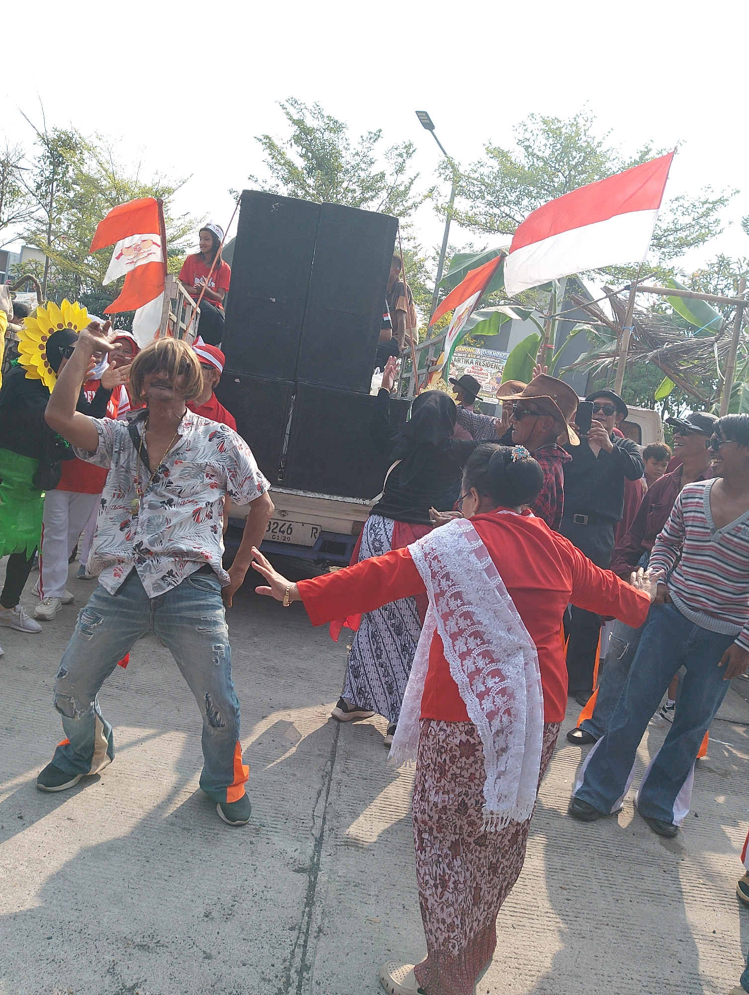 The image depicts a street scene in Karawang, Indonesia, where a group of people are gathered, seemingly celebrating or participating in an event. In the foreground, a man with a wig and a patterned shirt is dancing. He is wearing ripped jeans with orange fabric accents and dark sneakers. To his right, a woman in a red top and a floral skirt, adorned with a white lace shawl, is also dancing. Several other individuals are visible in the background, some holding Indonesian flags. Behind them, a large speaker system is mounted on a truck, suggesting a public gathering or parade. The environment appears to be an outdoor, paved area, possibly a street or plaza, with trees and a bright, sunny sky overhead. The presence of flags and the festive atmosphere suggest a patriotic or cultural celebration.