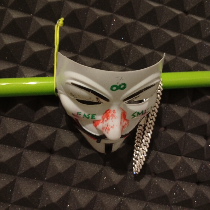 A white plastic mask depicting the face of the character "V" from the film "V for Vendetta" is lying on a surface covered in dark grey acoustic foam tiles. The mask has green lettering on it, reading "FASE" on the left side of the nose and "SMIL" on the right. The mask has a red-painted nose and features a chain-like accessory hanging from its right side. The mask is positioned in a way that it looks like it is wearing the chain. The mask is resting on a green pipe that has a yellow string attached to its upper left side. The location is Biedenkopf, Germany, which is a town in the Lahn-Dill-Kreis district in the state of Hesse, Germany. The coordinates are: latitude: 50.914, longitude: 8.465.