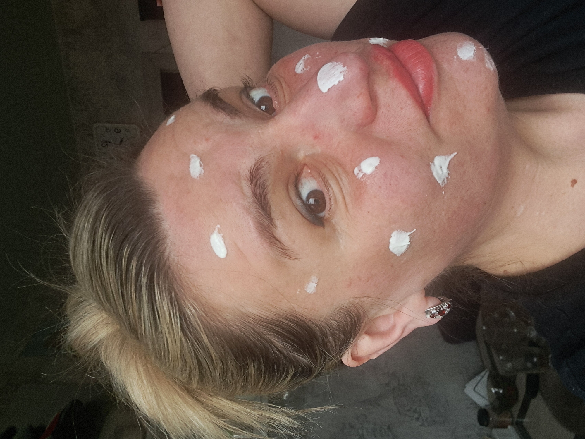 A woman with blonde hair pulled back in a low ponytail and red lipstick is looking upwards. Her skin has multiple white spots of cream or lotion on her forehead, cheeks, and neck. She is wearing a silver earring with red stones in her left ear. The background is blurry. The image was not taken in Elektrostal, Russia.