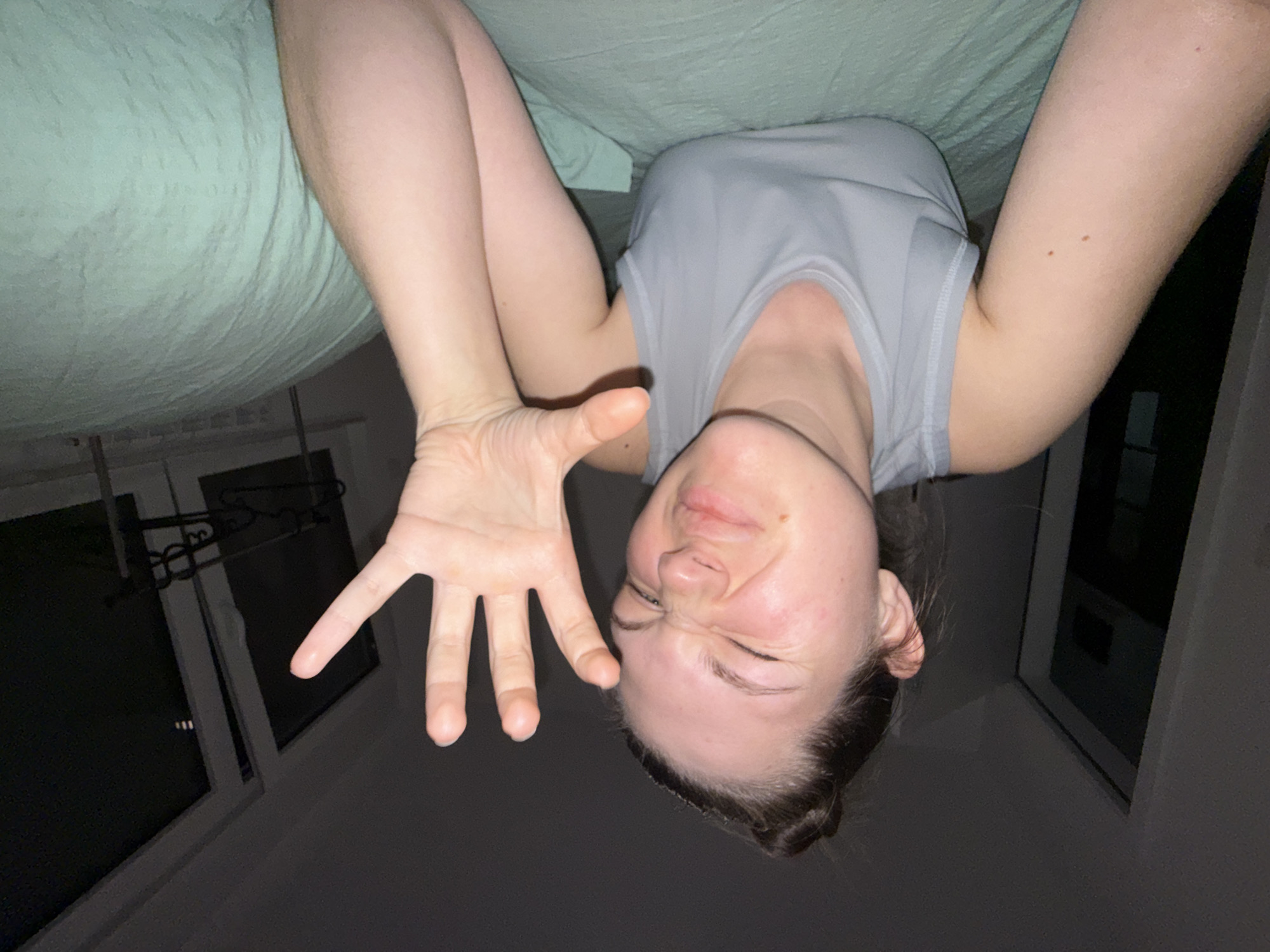 A woman is upside down, possibly on a bed or other soft surface, with her arms extended. She is wearing a grey sleeveless shirt. Her face is scrunched up as if in pain or discomfort. One of her hands is visible, with fingers spread open. A window is visible in the background. The location is not discernible within Vozdovac Urban Municipality, Serbia.