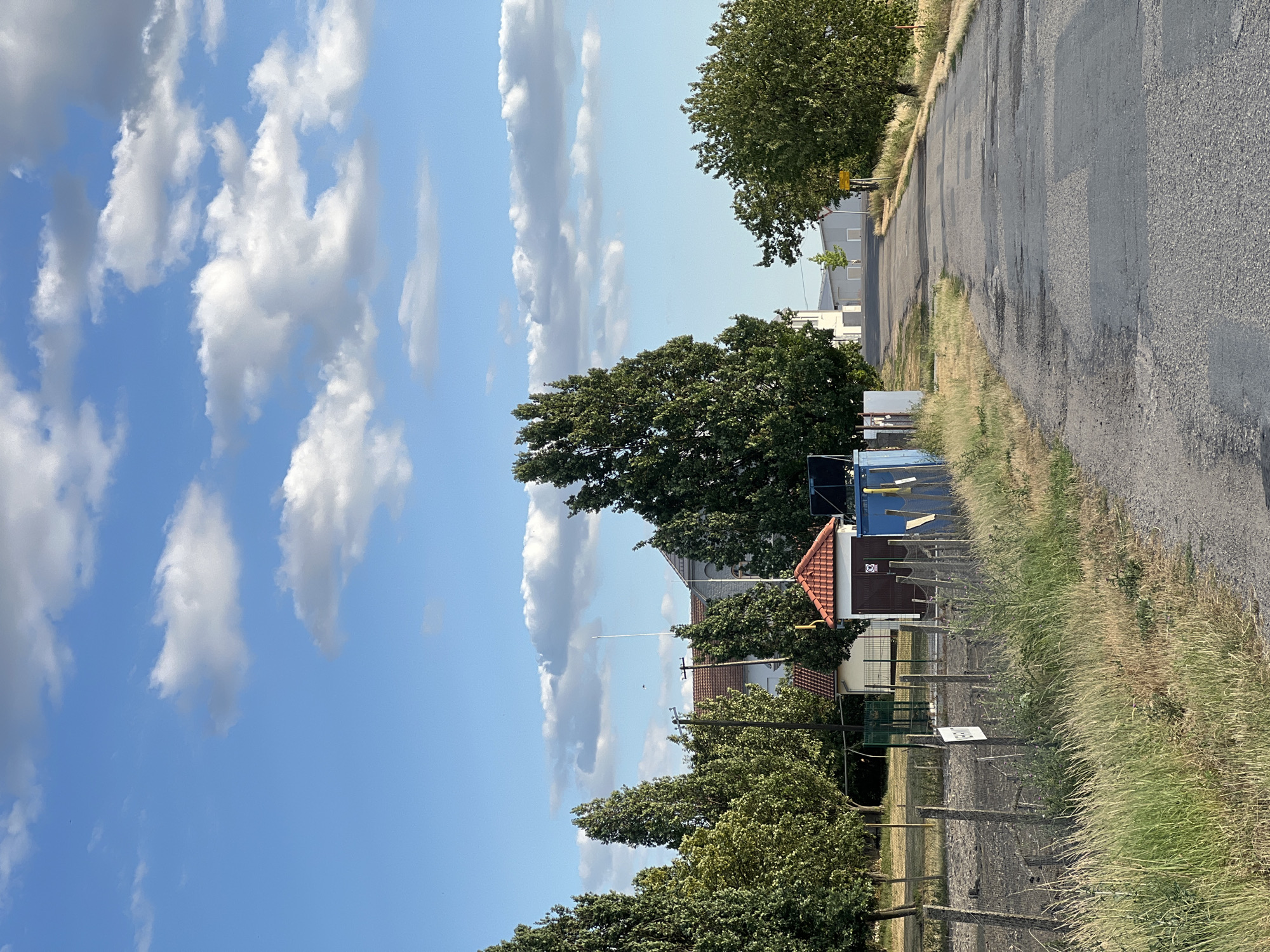 The image depicts a rural road leading through a grassy area with scattered buildings and trees under a blue sky with white clouds. The road is paved and shows signs of wear, with patches of darker asphalt. On the left side of the road, there is a fence made of wire and wooden posts, bordering an area with dry grass and some bushes. Beyond the fence, there are several trees, some tall and slender, others fuller. A small building with a red tiled roof and brown wooden doors is visible, possibly a shed or kiosk. A blue container is also present. In the background, there are larger buildings with white facades and tiled roofs, suggesting a residential or industrial area. On the right side of the road, more trees and dry vegetation are visible. The overall impression is of a quiet, sunlit day in a semi-rural setting, possibly on the outskirts of Žatčany, Czechia, given the context of the location. There are no people or animals visible in the image.
