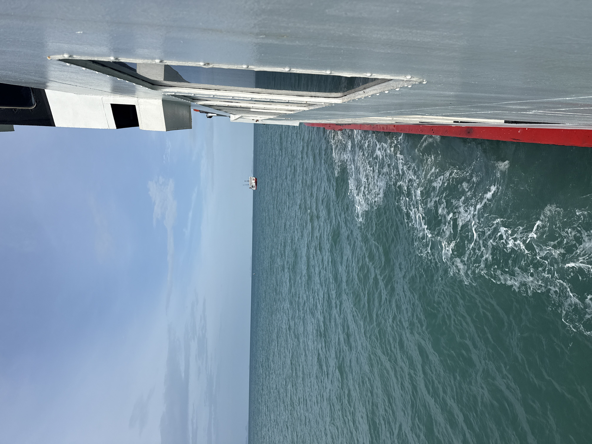 An exterior, full, eye-level shot depicts the side of a white ferry boat traveling through the choppy, teal ocean waters off the coast of Ko Samui District, Thailand. The ferry occupies the right half of the frame, its hull a stark white with a bright red stripe along its bottom edge. A line of rectangular windows with dark frames runs along the visible section of the boat's side. The water to the left of the ferry shows a foamy wake trailing behind the vessel, indicative of movement. In the distance, on the horizon where the ocean meets a pale blue sky with scattered clouds, a small white boat with a red accent is visible. The overall scene conveys a sense of travel and open sea.