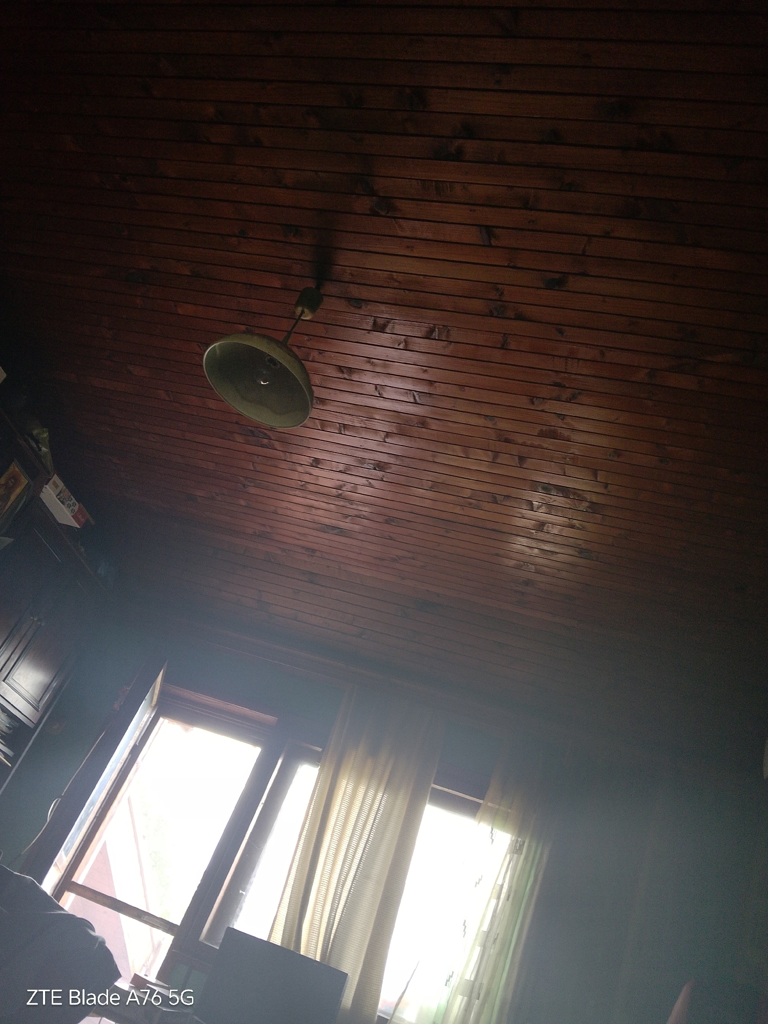The image shows the interior of a room with a wooden plank ceiling. A single light fixture hangs from the ceiling. Sunlight streams in through a window, illuminating sheer curtains. The lower part of the image shows furniture and a window with light filtering through. The bottom left corner of the image displays the text "ZTE Blade A76 5G". The location is the city of Stara Zagora, Bulgaria.