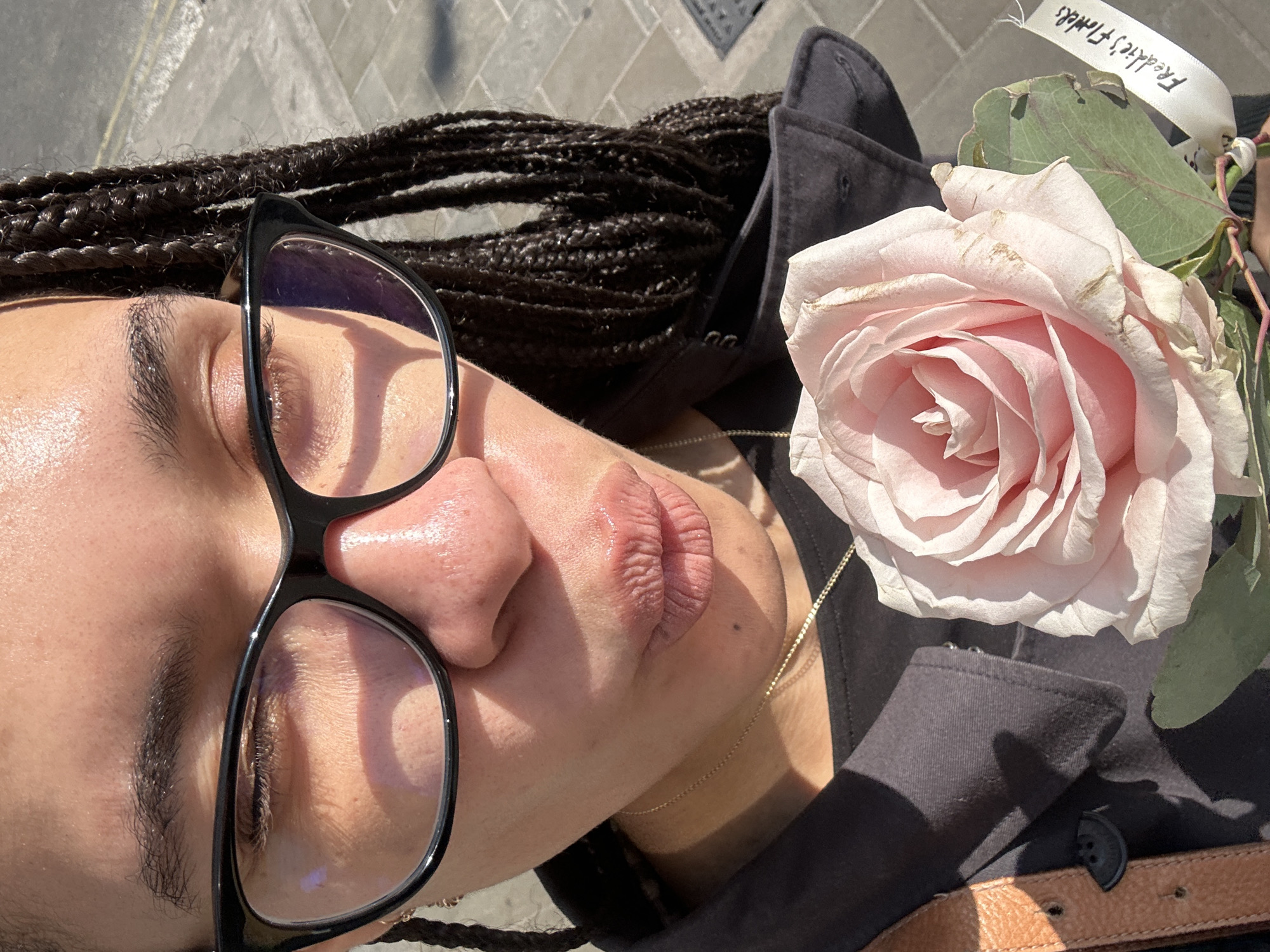A close-up image features a person lying down, with a pink rose held nearby. The person has brown braided hair and is wearing black-rimmed glasses. They appear to be fair-skinned. A thin gold necklace is visible around their neck. They are wearing a dark-colored jacket. The rose is light pink with some green leaves and a white ribbon attached that reads "Freddie's Flowers." The background appears to be a grey paved surface, possibly a sidewalk or street. The setting, based on the flower retailer mentioned on the ribbon and the architectural style of the urban environment, is likely London, United Kingdom. The perspective suggests the photo was taken from directly above the person lying down.