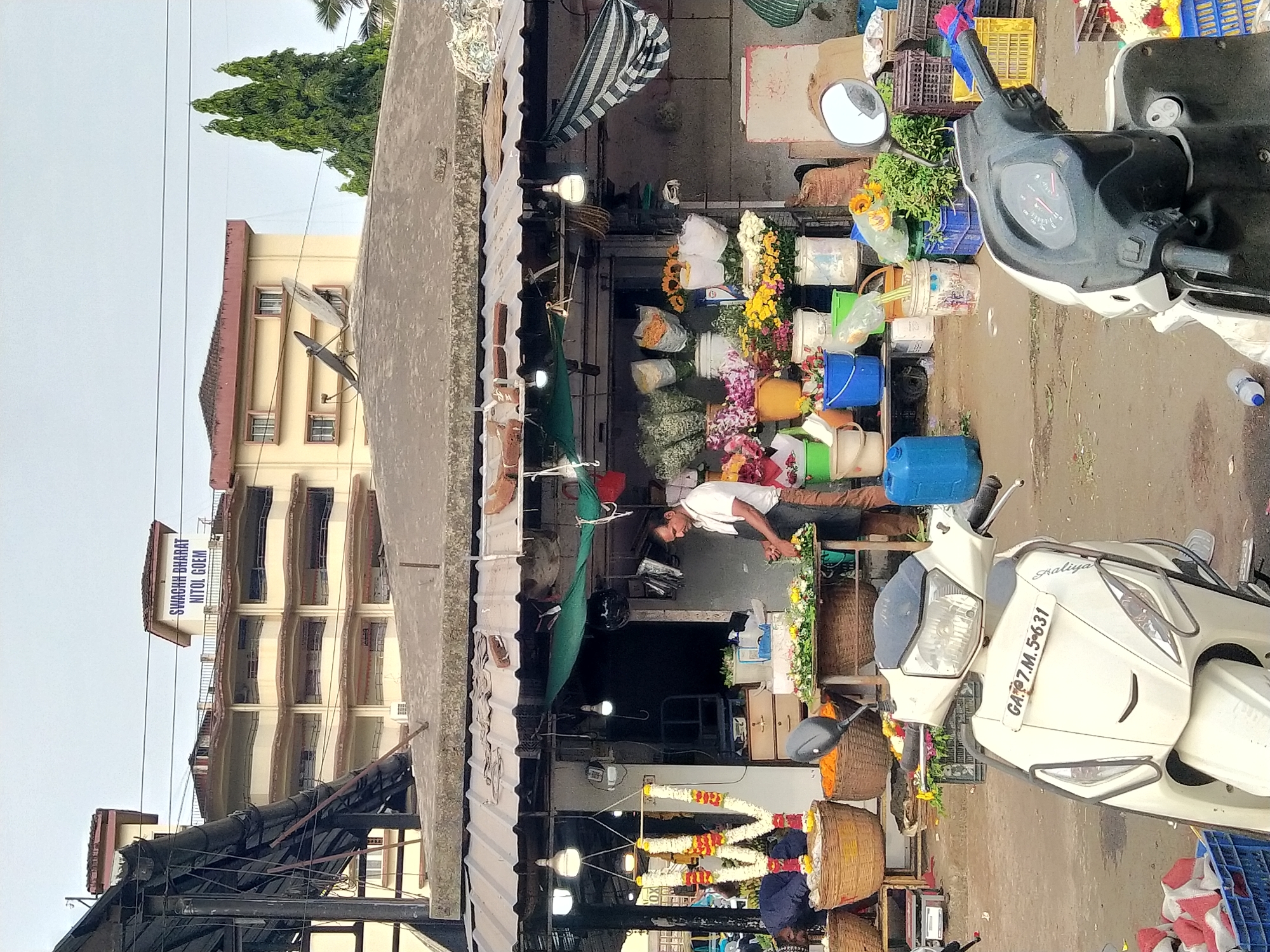 A street scene in Panaji, India, showcases a flower shop. A man wearing a white shirt and brown pants stands inside the shop, possibly arranging flowers or assisting a customer. The shop is stocked with various flowers, arranged in buckets and displayed in a colorful array. Garlands are also on display in the front of the shop. Outside the shop, two scooters are parked, one is a white scooter with the license plate GA-07-M-5631, and the other is gray and black. In the background, a multi-story building with balconies can be seen with “SWAGRHA BHARAT NITOL GOEM” written on top.