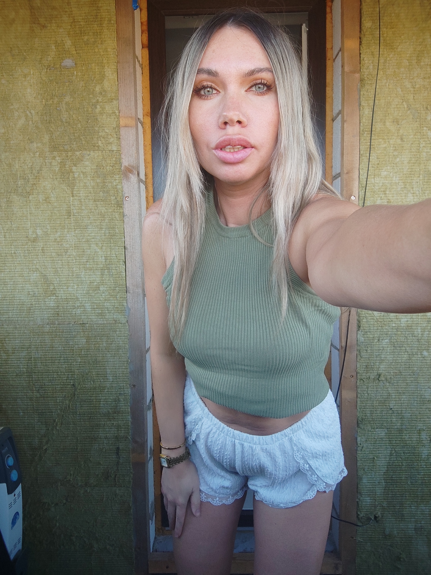 A woman stands in a doorway, taking a selfie. She has blonde hair with dark roots, light skin, and light-colored eyes. She wears a light green ribbed tank top and white lace shorts. A gold watch is visible on her left wrist. The doorway is framed by wood, and behind the woman is what appears to be insulation material. A section of wall is visible to the left of the doorway, also made of the same insulation material. Part of what looks like an appliance or electrical box is visible in the lower left corner. The location of Votkinsk, Russia, cannot be confirmed from the image.