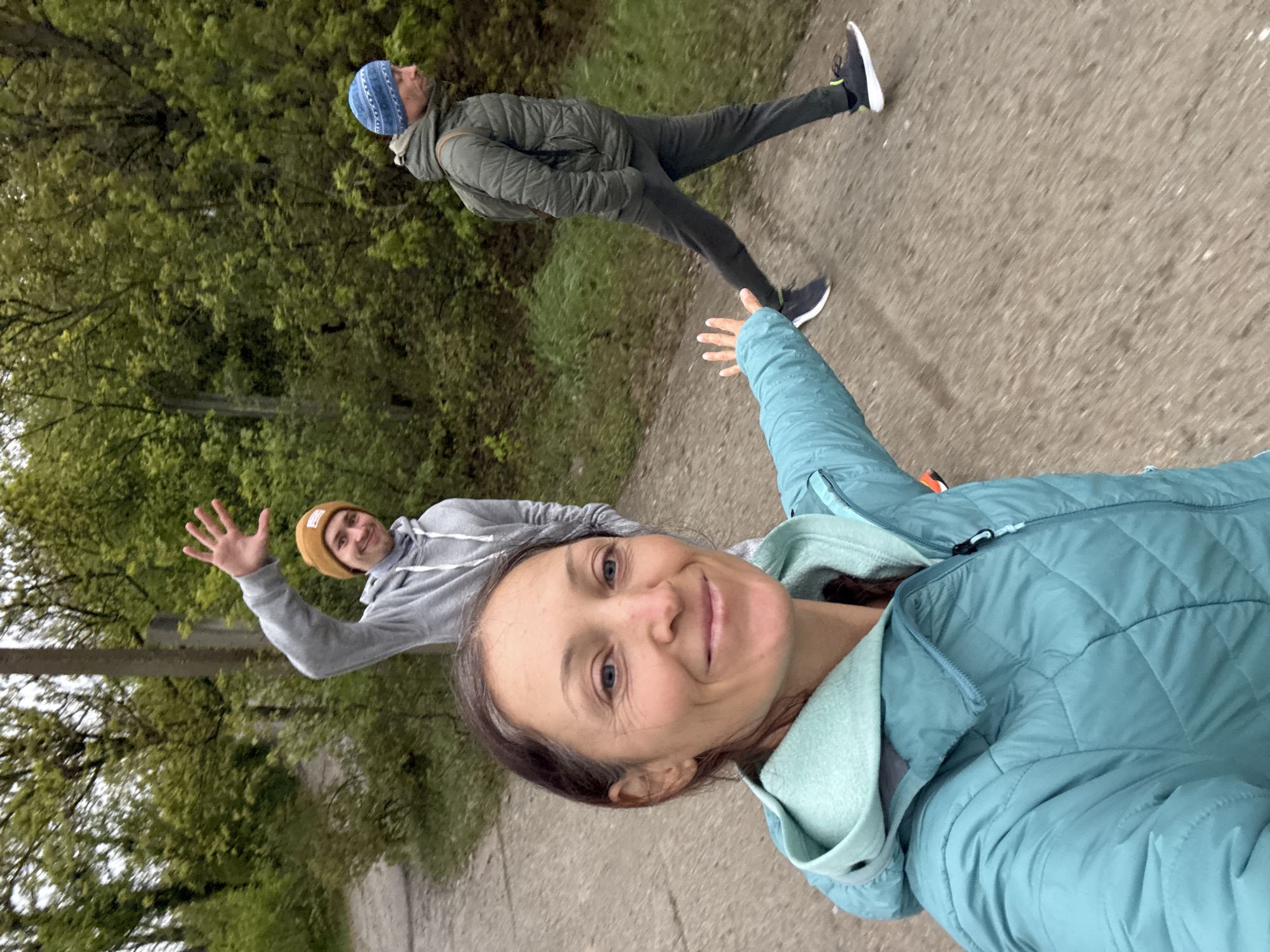 Three people are walking on a paved path bordered by trees, likely in Kiparisnoe, Ukraine, although specific urban features are not visible. A woman in the foreground is taking a selfie, smiling and extending her arm toward the camera. She wears a light blue jacket and has dark hair. Behind her to the left is a man wearing a gray hoodie and a tan beanie, waving. Further behind and to the right is another man in a green jacket and blue beanie. The path and surrounding greenery suggest an outdoor recreational area.