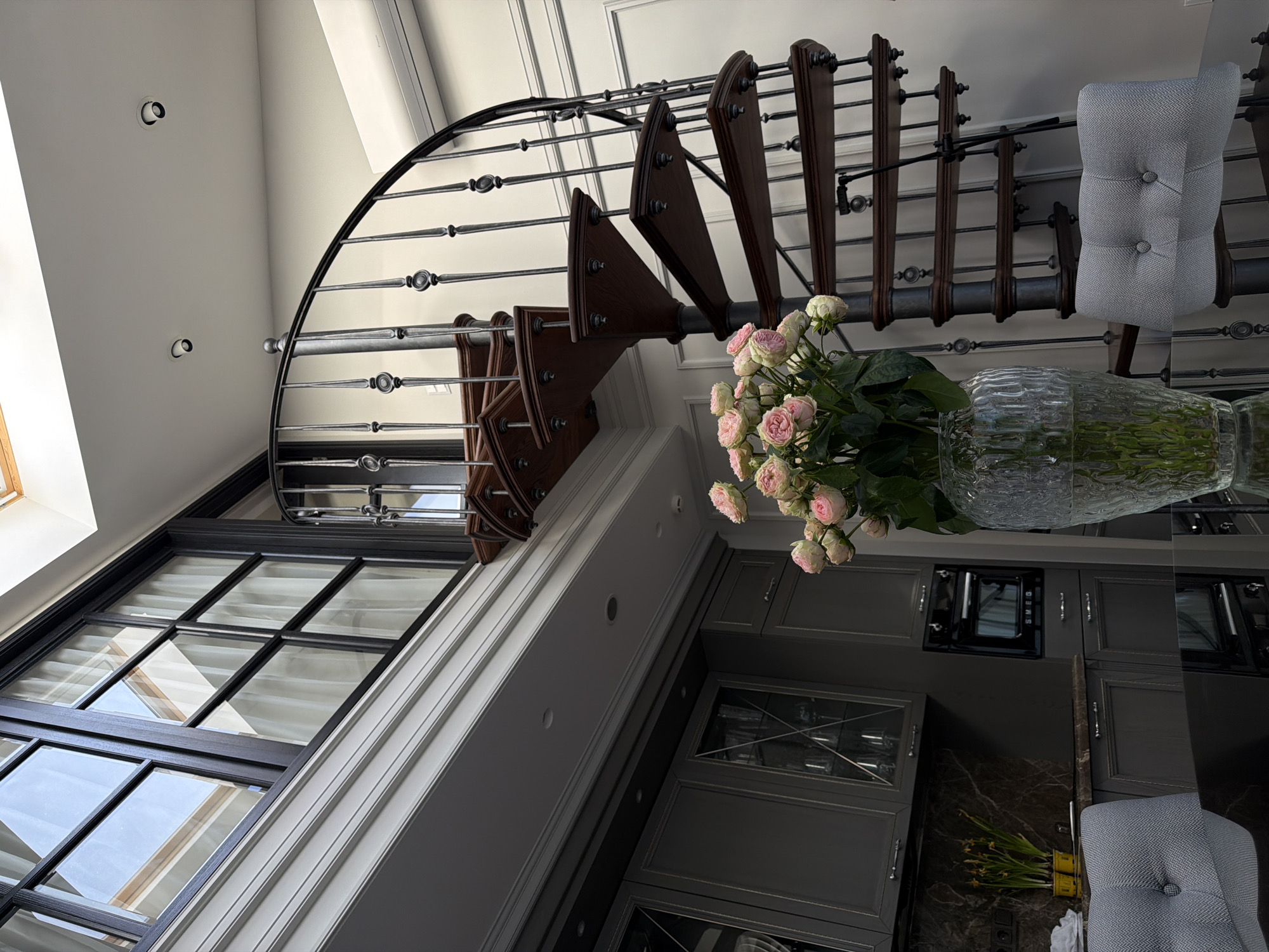 The image depicts the interior of a multi-level dwelling in Moscow, Russia. The scene focuses on a dining area and kitchen integrated into a space featuring a prominent spiral staircase. In the foreground, a reflective black dining table is partially visible, hosting a clear glass vase containing a bouquet of pink and white roses. Two light grey tufted dining chairs with a herringbone pattern are positioned at the table. To the left, a kitchen area features dark grey cabinetry, a dark marble backsplash, and an integrated black SMEG oven. Yellow potted plants rest on the kitchen counter. Dominating the right side is a dark brown wooden spiral staircase with a black wrought iron railing, leading upwards to a second level. The upper level features large black-framed windows with white curtains and a white wall with decorative molding. The ceiling is white with recessed lighting fixtures and a visible skylight. A white wall-mounted air conditioning unit is positioned on the wall near the staircase. No people are present in the scene.