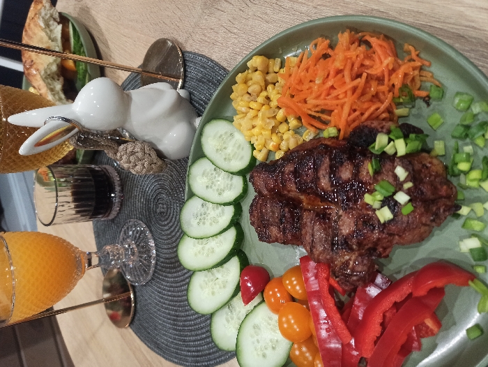 A close-up, elevated view depicts a prepared meal set on a light-colored wooden table in Nizhneye Zadolgoye, Russia. The primary subject is a matte green-grey plate presenting a grilled piece of meat, exhibiting distinct grill marks and a glossy dark brown surface, topped with chopped green onions. Arranged around the meat are several fresh components: eight circular slices of cucumber, one red cherry tomato and approximately nine orange cherry tomatoes, multiple longitudinal strips of red bell pepper, a portion of whole kernel corn, and a pile of finely shredded orange carrots. Additional chopped green onions are scattered across the plate. In the background, positioned on a circular dark grey placemat with a woven texture, are three beverages: a clear ornate glass goblet filled with orange juice, a small dark ribbed glass containing a dark liquid, and a partially visible clear goblet with an amber beverage. A small white ceramic rabbit figurine, featuring a gold-colored accent and a small woven brown accessory attached to its back, stands on the placemat. A blurred green plate with a partially visible food item, resembling a sandwich or pastry, is also present in the background. No people are visible in the scene.