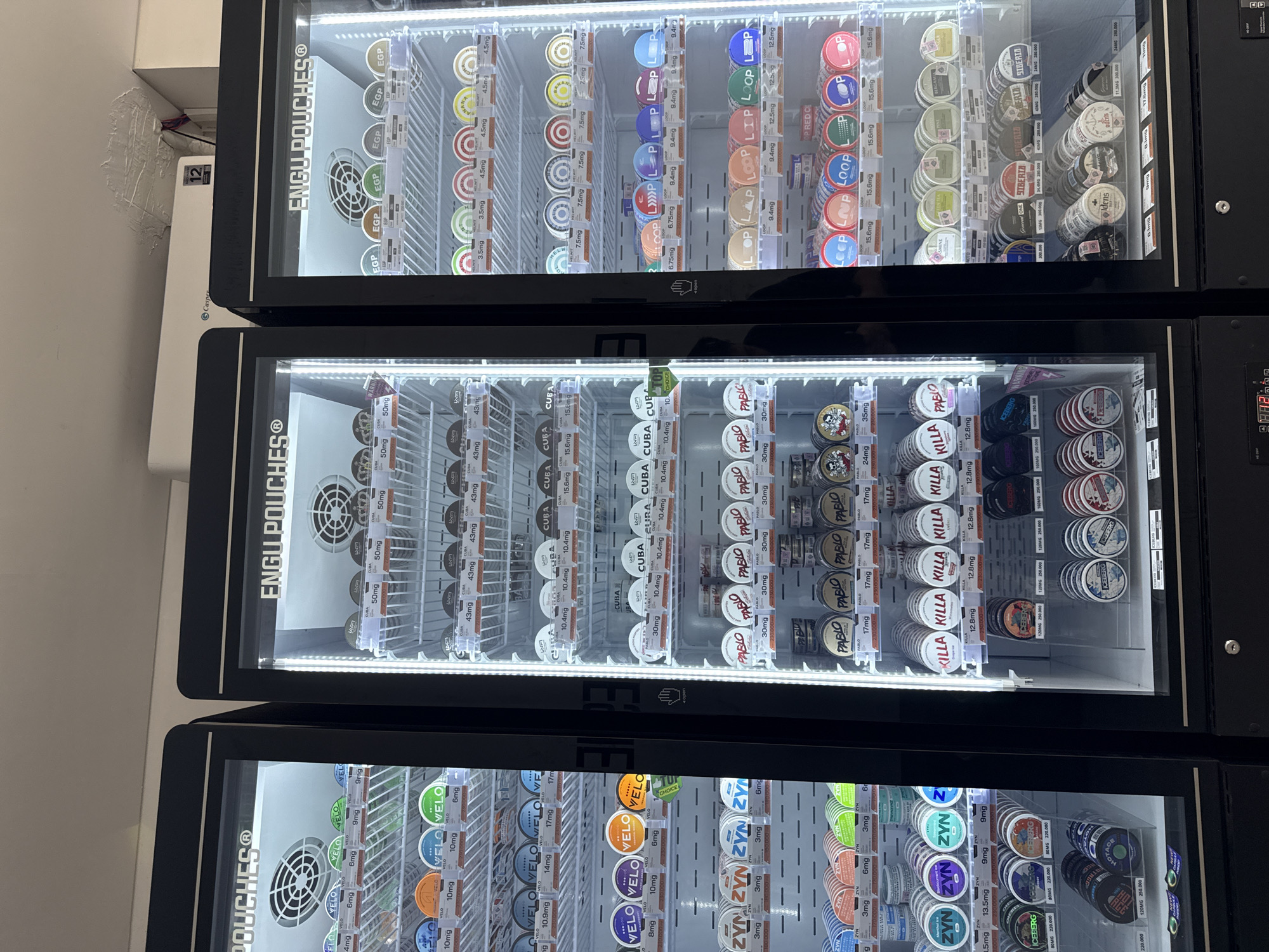 The image displays three stacked vending machines filled with cylindrical cans of nicotine pouches. The vending machines are black with transparent fronts, allowing a clear view of the contents inside. Each can is labeled with its respective brand and nicotine content, including brands like "ENGU POUCHES," "VELO," "ZYN," "CUBA," "PABLO," "KILLA," and "ICEBERG." The text in the picture indicates different nicotine levels in each product. The background is a plain white wall. It is unclear from the picture whether this is in Đà Nẵng, Vietnam.