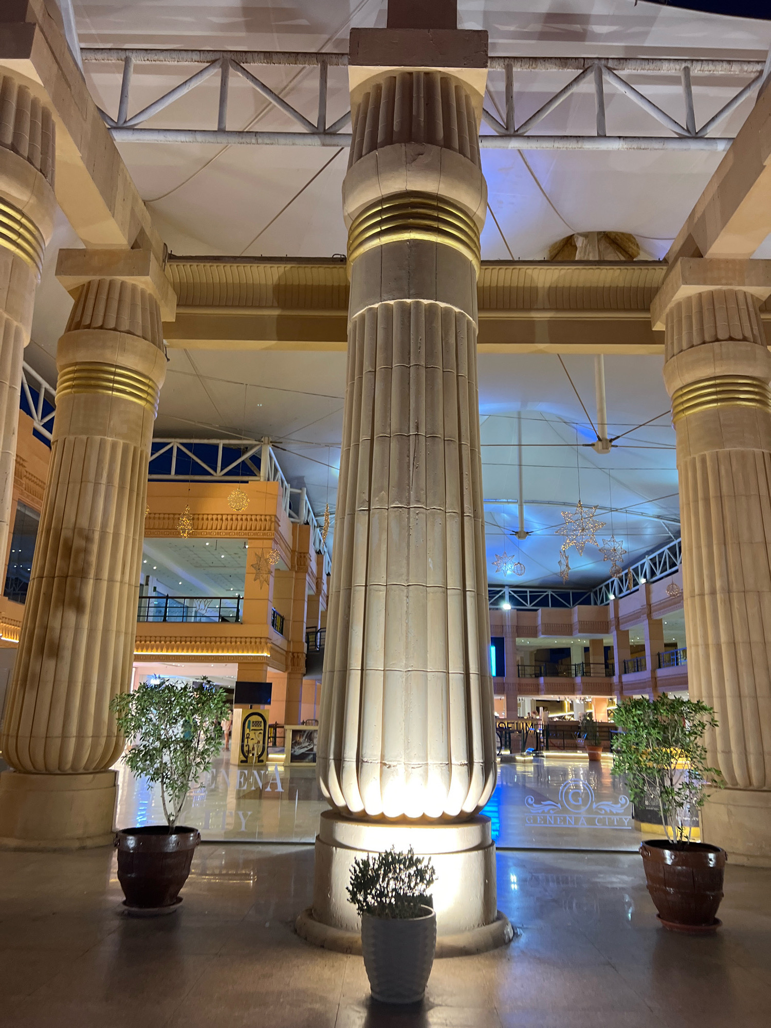 The image depicts the interior of a shopping mall in Sharm El Sheikh, Egypt, designed with an Egyptian architectural theme. Dominating the foreground are several large, fluted columns with gold accents, reminiscent of ancient Egyptian temples. These columns support a high, modern ceiling with exposed structural elements and a translucent fabric covering.
In the midground, multiple levels of the mall are visible, featuring balconies, railings, and storefronts. Decorative hanging light fixtures, some resembling stars, are suspended from the ceiling, adding to the ambiance. Blue lighting illuminates parts of the interior, creating a striking contrast with the warm tones of the columns and building facade.
In the foreground, two potted plants add a touch of greenery to the scene. A large plant in a dark brown, barrel-shaped pot is positioned to the left of the central column, while a smaller, bushier plant in a grey pot sits in front of the right column.
The floor appears to be polished and reflective, mirroring the lights and architectural features. A glass partition in the midground displays the text "GENENA CITY" in white lettering, indicating the name of the mall. No people are clearly visible or interacting in the frame. The overall impression is one of grandeur and a unique blend of historical inspiration and modern retail design.