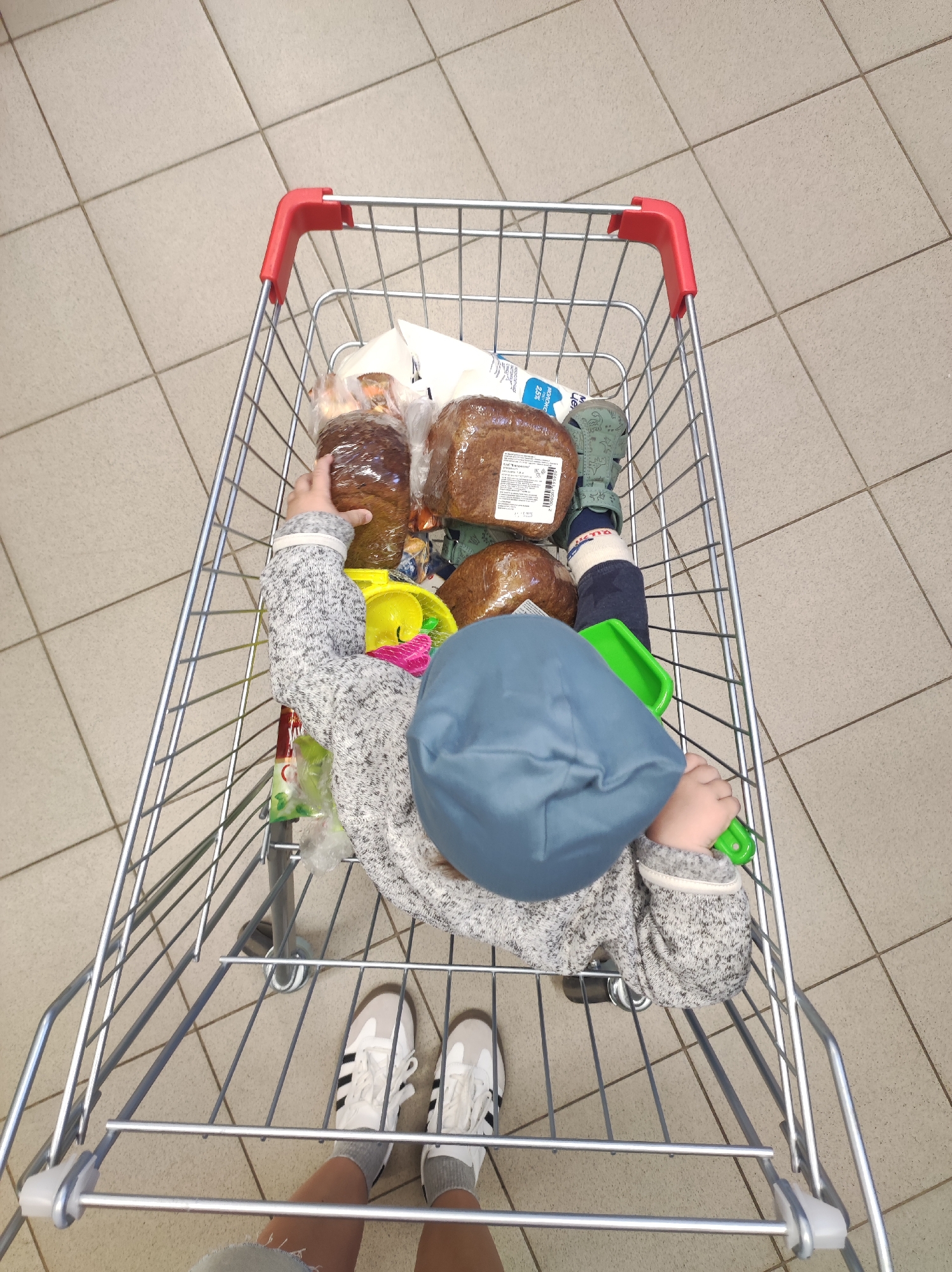 A first-person perspective image depicts a shopping cart in a store, possibly in Chaikovsky, Russia, given the presence of Russian-labeled bread. Inside the cart is a young child, wearing a blue hat and a gray sweater. There are several loaves of dark bread and plastic toys surrounding the child. The cart is on a tiled floor. A person's legs and sneakers are visible at the bottom of the image, presumably the person pushing the cart.
