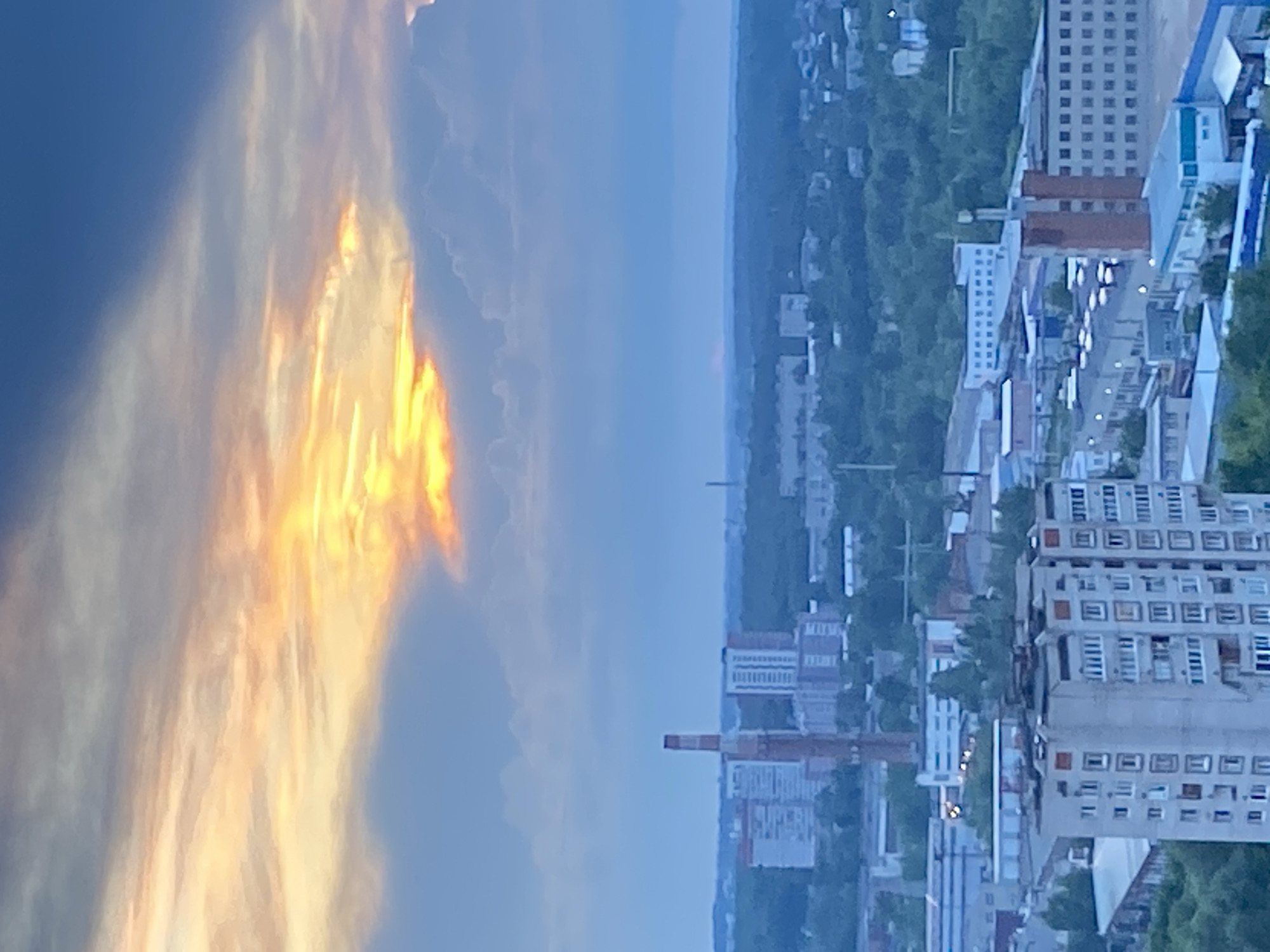 The image captures an aerial view of a cityscape, likely Izhevsk, Russia, during either sunrise or sunset, as indicated by the dramatic interplay of light and clouds. The sky is dominated by a brilliant orange and yellow glow emanating from behind a mass of pale blue and white clouds, creating a striking contrast against the deeper blue sky above and around.
Below the sky, a dense urban and industrial landscape unfolds. Prominent features include several multi-story residential buildings and industrial structures. A tall, red and white striped smokestack or industrial chimney stands out in the mid-ground. Numerous factories and smaller buildings are scattered throughout the scene, interspersed with lush green trees. The overall impression is one of a sprawling industrial city under a dynamic sky. No people are visible in the image.