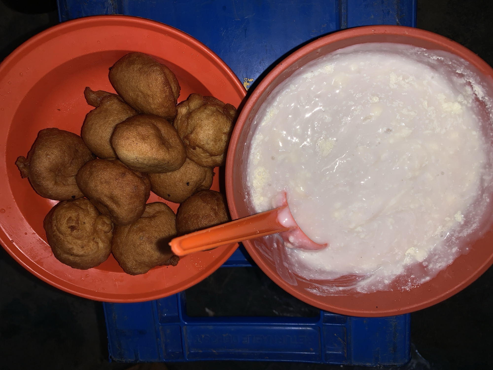 The image presents a top-down view of two orange plastic bowls filled with food, resting on a blue plastic surface, likely a crate or stool. The scene appears to be set in a casual environment, potentially at night or in low light, given the dark background and the way the food is illuminated.
In the left bowl, there is a pile of golden-brown, irregularly shaped fried dough balls. These appear to be a popular snack such as puff-puff, a common street food in Nigeria. The surface of the dough balls is slightly textured and some exhibit small folds or creases from the frying process.
The right bowl contains a thick, creamy white substance, which could be pap (ogi) or a similar fermented cereal pudding, often consumed in Nigeria. An orange plastic spoon is partially submerged in this creamy mixture, suggesting it is ready to be eaten. The texture of the pap shows some minor clumps or undissolved bits, characteristic of its preparation.
The blue surface beneath the bowls is a plastic object, possibly a crate, with visible grid-like patterns and some faint, blurry white text on its side. The text is largely unreadable, but segments like "LETU" and "MJE" are vaguely discernible. The presence of these food items and the informal setting are consistent with local culinary practices in regions like New Stadium, Nigeria. The overall impression is a simple, traditional meal or snack.