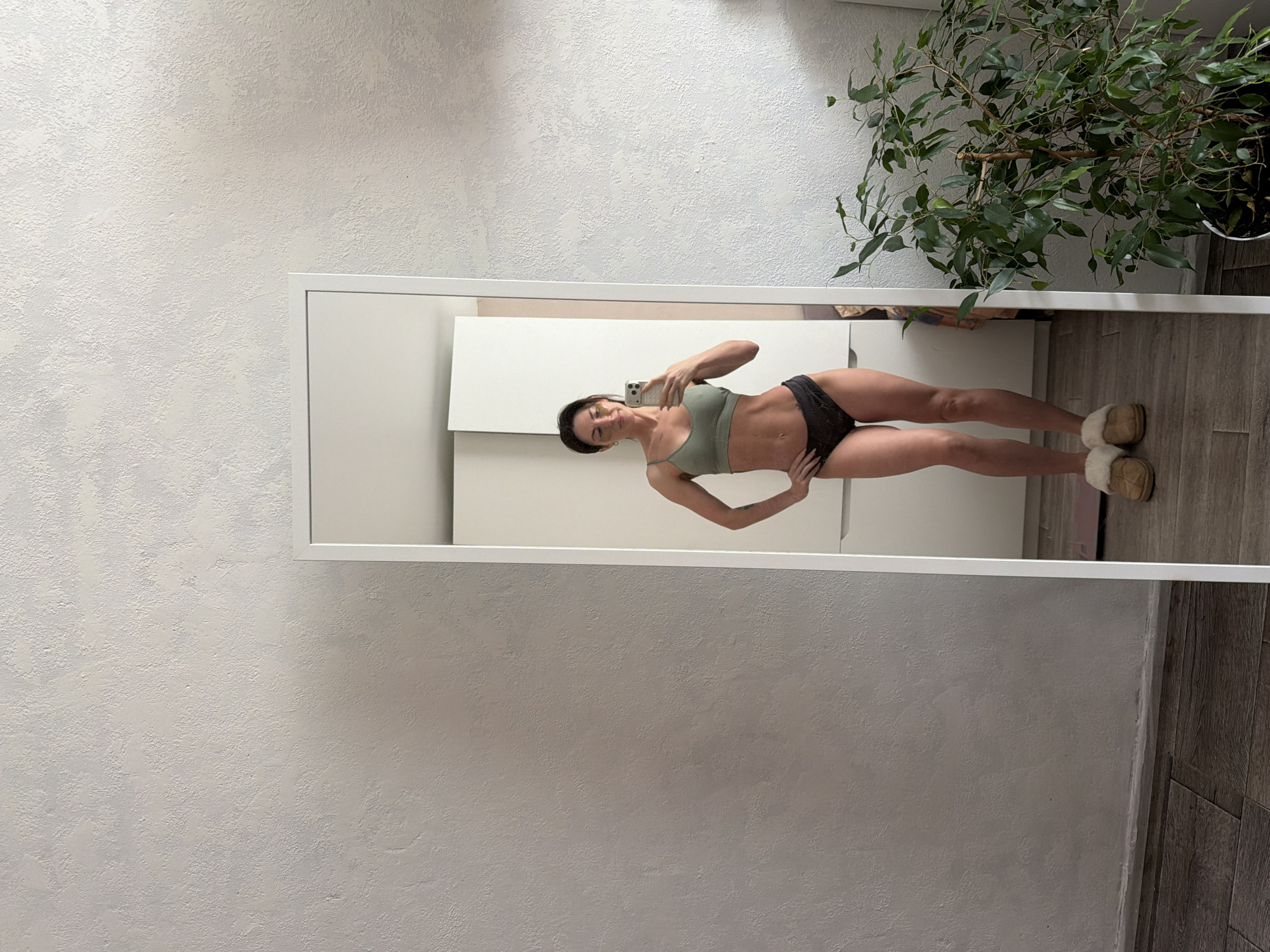 An indoor mirror selfie captures a woman posing in what appears to be a residential setting in Perm, Russia. The woman, with dark hair, is wearing a sage green sports bra or bikini top and dark briefs. She holds a light-colored smartphone with her right hand to take the picture, while her left hand rests on her hip. She has a subtle smile or relaxed expression.
The reflection shows a brightly lit room with light-colored, textured walls, possibly stucco. To the right of the mirror, a large potted plant with abundant green leaves, resembling a ficus, is visible. The floor in the reflection appears to be wood-look laminate or tile. Behind the woman, within the mirror's frame, there are simple white furniture pieces, possibly cabinets or a dresser. The woman is also wearing fuzzy, light brown slippers. The overall impression is a modern, minimalist interior. No specific time of day or weather conditions are discernible, but the lighting suggests daytime. There is no visible text in the image.