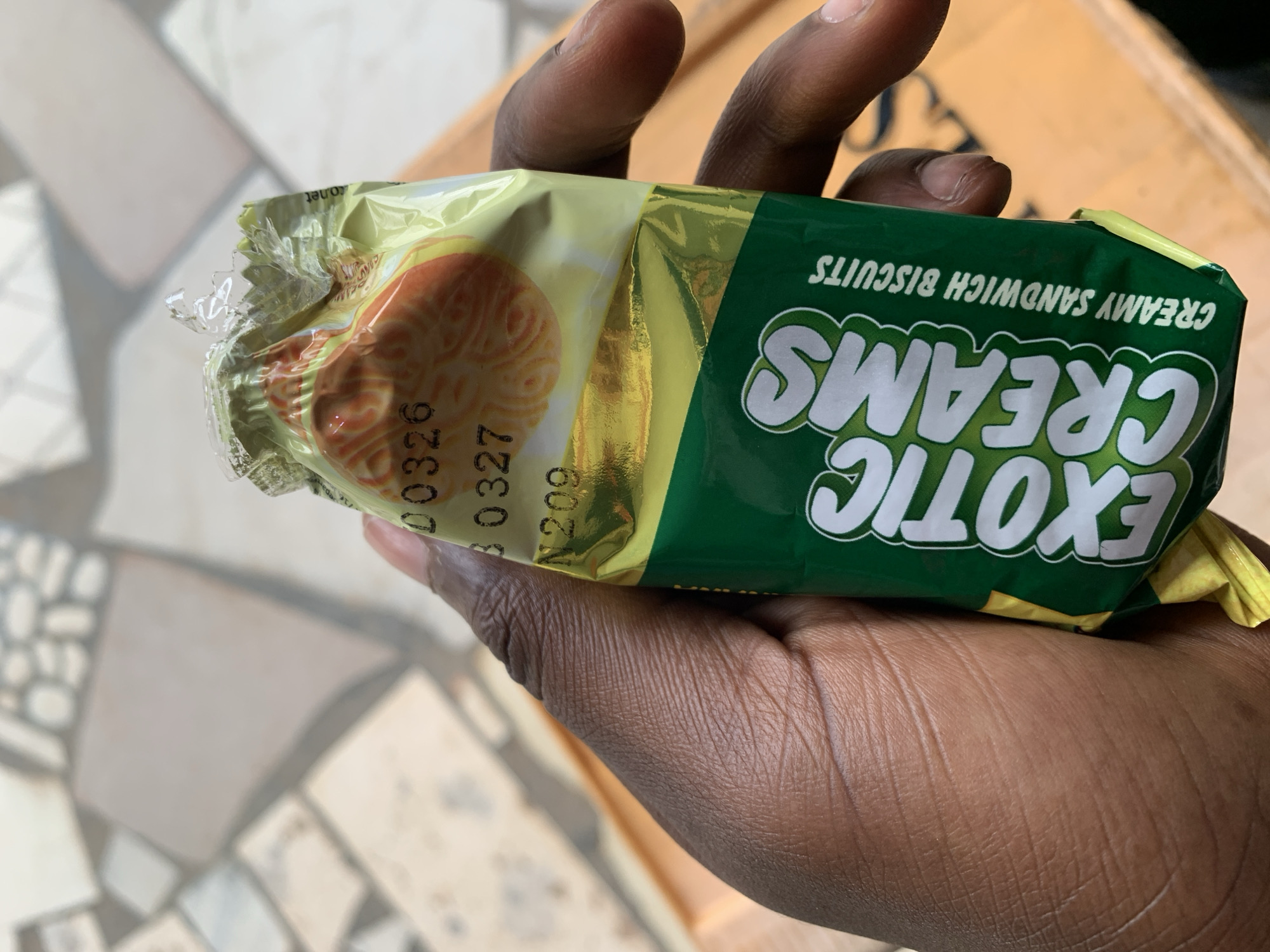 A person is holding a packet of "Exotic Creams" creamy sandwich biscuits in their hand. The packet is green and yellow, with an image of a biscuit on the wrapper. Part of the wrapper has been torn off. The location is unknown, but the city is New Stadium, Nigeria. The background shows a tiled floor and a cardboard box.