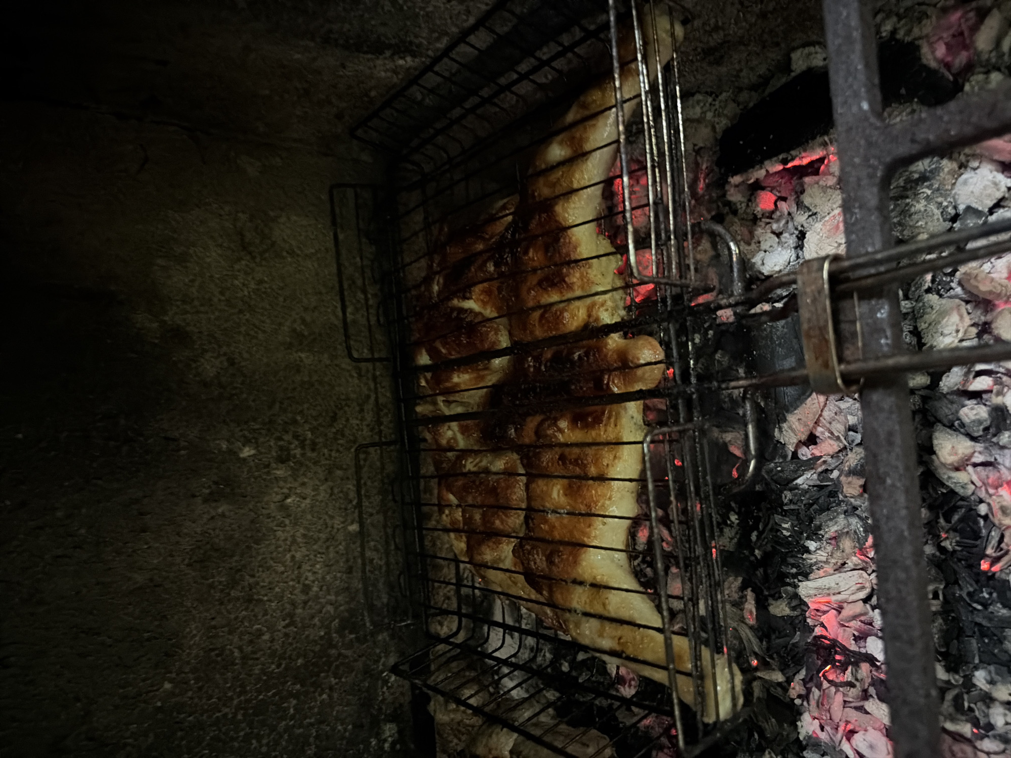The image shows food, likely chicken, being cooked on a grill over hot coals. The food appears golden brown and is positioned within a metal grilling basket. The background is dark and appears to be the inside of a grill or oven, with a rough, stone-like texture on the upper walls. Red embers glow from the coals beneath the grill. No people are visible in the image. The location is the city of Balashikha, Russia.