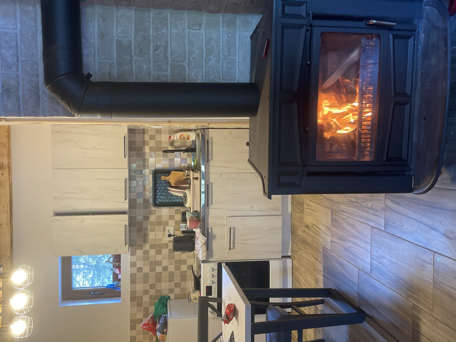 This image depicts an interior scene in Palež, Montenegro, featuring a rustic kitchen and an active wood-burning stove.
In the foreground, a dark, cast-iron wood-burning stove is prominently positioned on the right, with visible orange flames and burning wood through its glass door. A dark metal chimney pipe ascends from the stove, bending 90 degrees to the right and connecting to a light-colored brick wall. A small, red implement rests on the top surface of the stove. The floor consists of rectangular tiles with a light wood-grain pattern.
To the left of the stove, a light-colored table with dark metal legs is partially visible, with a dark chair tucked underneath. A crumpled red wrapper or bag lies on the table surface.
The background reveals a kitchen area. Light-toned wooden cabinetry, comprising both base and wall units with dark handles, lines the back wall. A stainless steel sink with a black faucet is present, flanked by a dish drying rack containing bowls and a cutting board, and a bottle of Fairy-brand dish soap. Various kitchen items, including a black kettle and a small white pot, are on the countertop. A beige and brown checkered tile backsplash covers the wall above the countertop. To the far left, a white refrigerator stands next to a white electric or gas stove with a frying pan on one burner. Above the upper cabinets, a small window offers a view of green foliage bathed in daylight. Exposed wooden beams are visible on the ceiling. No individuals are present in the scene.