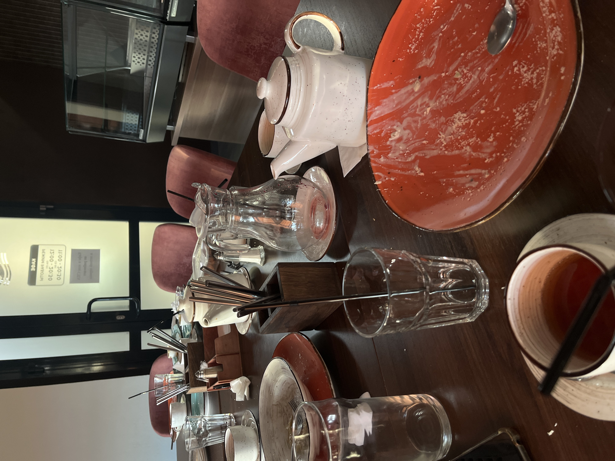 The image depicts a table cluttered with dirty dishes and tableware in what appears to be a restaurant or cafe. The location is likely Chaikovsky, Russia, based on a sign visible through a glass door indicating business hours in Russian (13:00-20:30). The table is wooden and covered with a used orange plate, a white teapot, a glass pitcher, several glasses, a mug with tea, a container filled with metal straws, and miscellaneous cutlery. Crumbs and food residue are visible on the plate. The scene suggests a meal has been consumed. Several pink chairs are visible around the table. Through the glass door, the outside world can be seen.