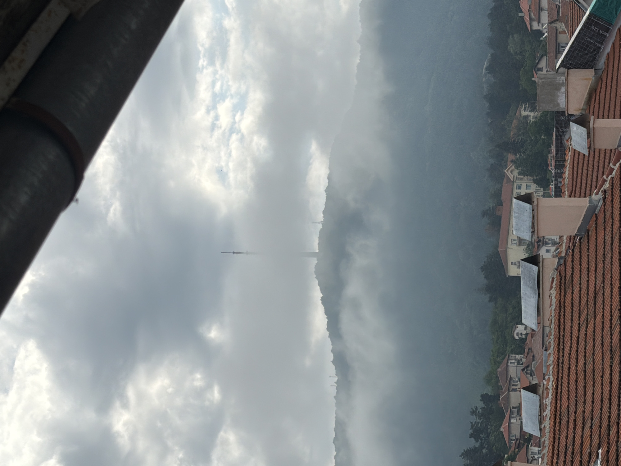 The image shows a view of the city of Belogradchik, Bulgaria, from a rooftop. In the foreground, there are several red-tiled roofs and chimneys of residential buildings. In the middle ground, there are more buildings with different roof types and colors, along with some trees. The background features a large, forested hill shrouded in mist, with a tall communication tower visible at its peak. The sky is overcast with thick, grey clouds. The scene suggests a damp or foggy day.