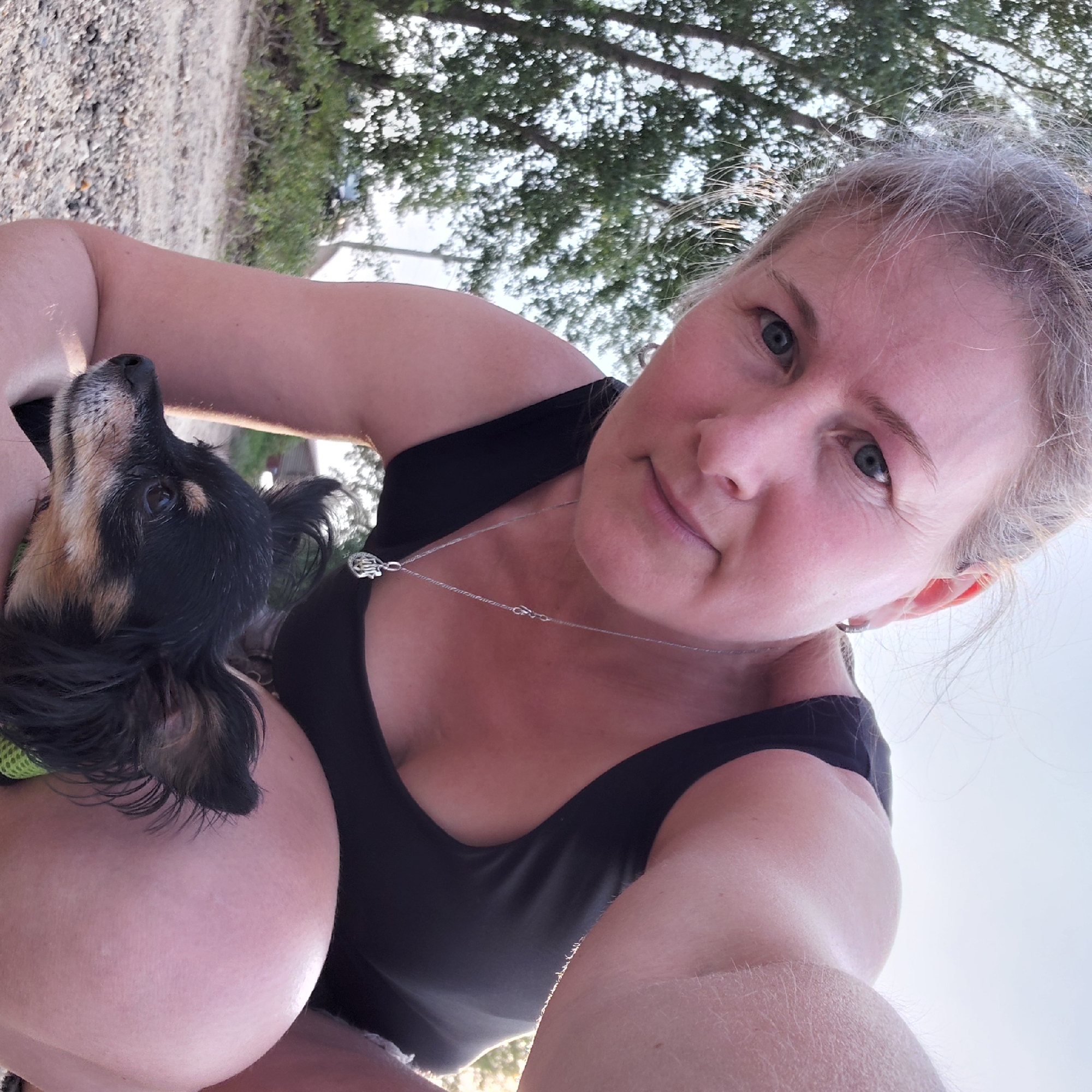 The image is a selfie taken outdoors, likely during daytime, given the bright lighting. A woman with blonde hair pulled back and visible facial features, including blue eyes and a slight smile, is crouching. She is wearing a black top and a silver necklace with a pendant. A small black and tan dog is being held in her arms, its head visible in profile. The background shows trees and a gravelly or sandy ground, suggesting a park or natural area. The image quality is slightly grainy. The location is identified as Novosibirsk, Russia.
