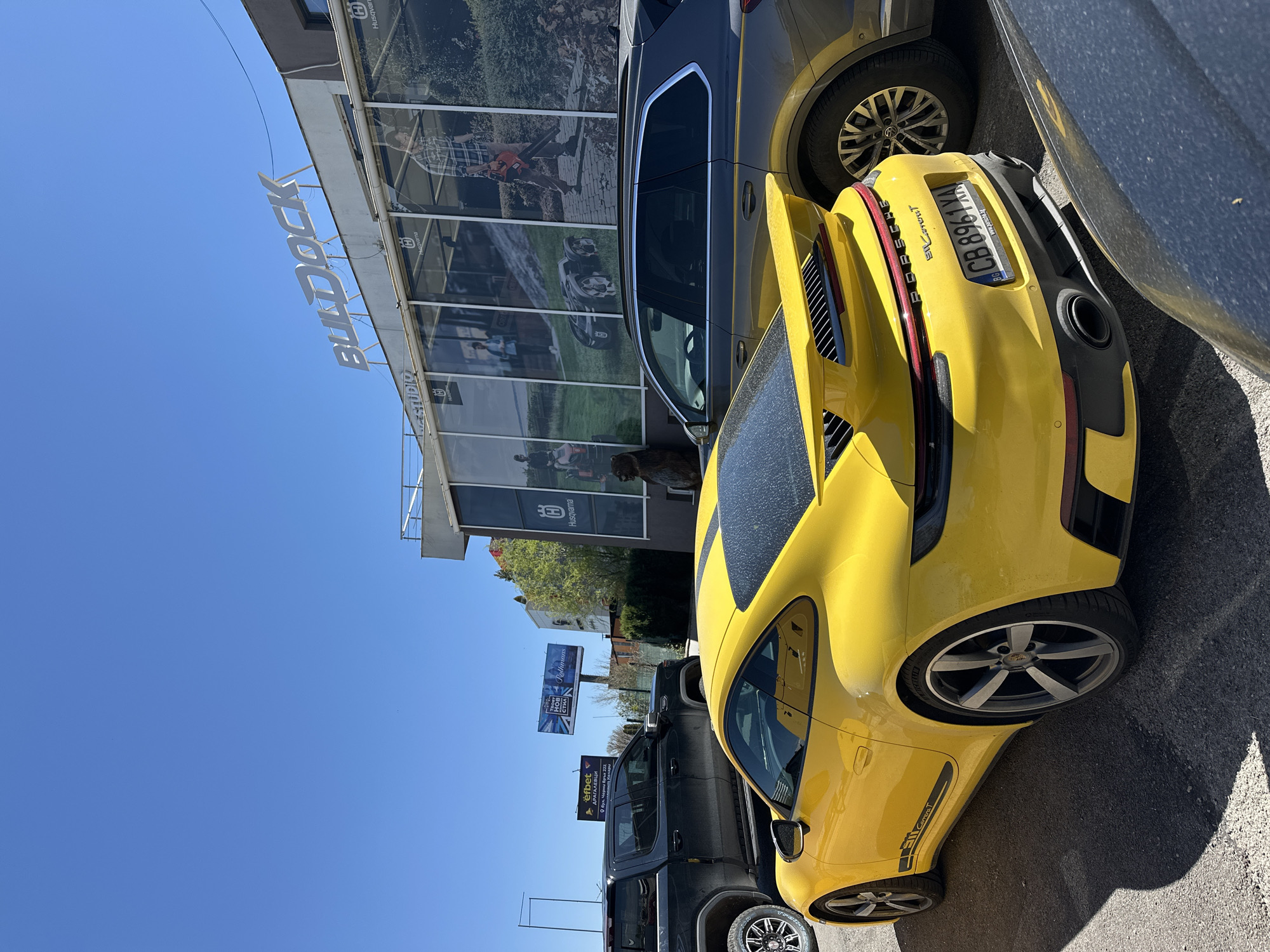 The image shows a yellow Porsche Carrera T parked on a street in Sofia, Bulgaria. The car has license plate "CB 8961 XA", and the text "Ellarorut" is present on the back. It's parked near a building with "BULDOCK STUDIO" and "Husqvarna" signage, with associated imagery and logos in the windows. A gray vehicle is parked to the left of the Porsche. In the background, there are billboards, including an "efbet" advertisement, and "Rollmans". The location appears to be in the Dragalevtsi district of Sofia, as indicated by the "efbet ДРАГАЛЕВЦИ" sign. The sky is clear and blue. There are other vehicles partially visible, including a gray SUV to the left of the Porsche and a dark-colored pickup truck further in the background.