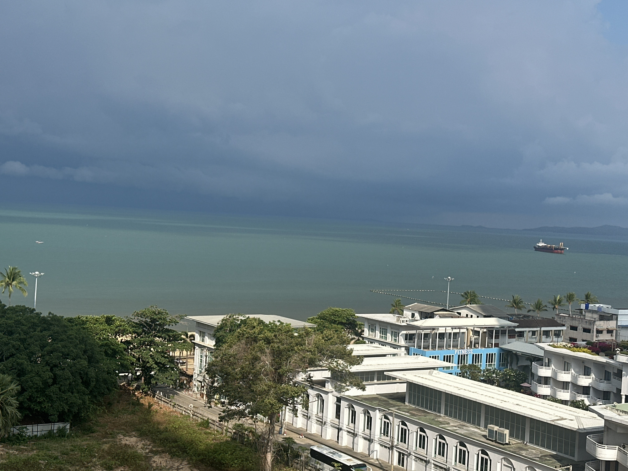 This high-angle, wide shot captures a coastal city merging with the sea under a dramatic, overcast sky. The scene is set in what appears to be a bustling urban area alongside a calm ocean.
In the foreground, numerous white and light-colored buildings with distinct architectural features, including balconies and arched windows, dot the landscape. Many green trees, including palm trees, are interspersed among the structures and line the coast, suggesting a tropical environment. A road is visible winding through the buildings, and the top of a bus can be seen on it. A tall street light pole stands on the left.
The middle ground is dominated by the expansive, greenish-blue sea, which appears calm with only faint ripples on its surface. Near the shore, a line of buoys or a netted area is visible, possibly demarcating a swimming or mooring zone. Further out in the sea, a large cargo ship or tanker with a red hull and a brown superstructure is either anchored or moving slowly towards the right. A smaller, lighter-colored vessel is also visible closer to the left side of the sea.
The background is defined by a heavy, dark grey, and heavily clouded sky that spans across the entire horizon, indicating impending rain or stormy weather. Despite the dark clouds, some lighter patches are visible higher up. Distant landmasses, possibly hills or islands, can be seen faintly on the horizon to the far right.
A notable detail is the word "FITNESS" written on the side of a prominent blue building in the mid-right section of the city. The overall atmosphere conveyed by the dark sky is one of impending weather change, giving the scene a somber yet dramatic feel. The architecture and vegetation cues align with a coastal resort city like Pattaya, Thailand.
