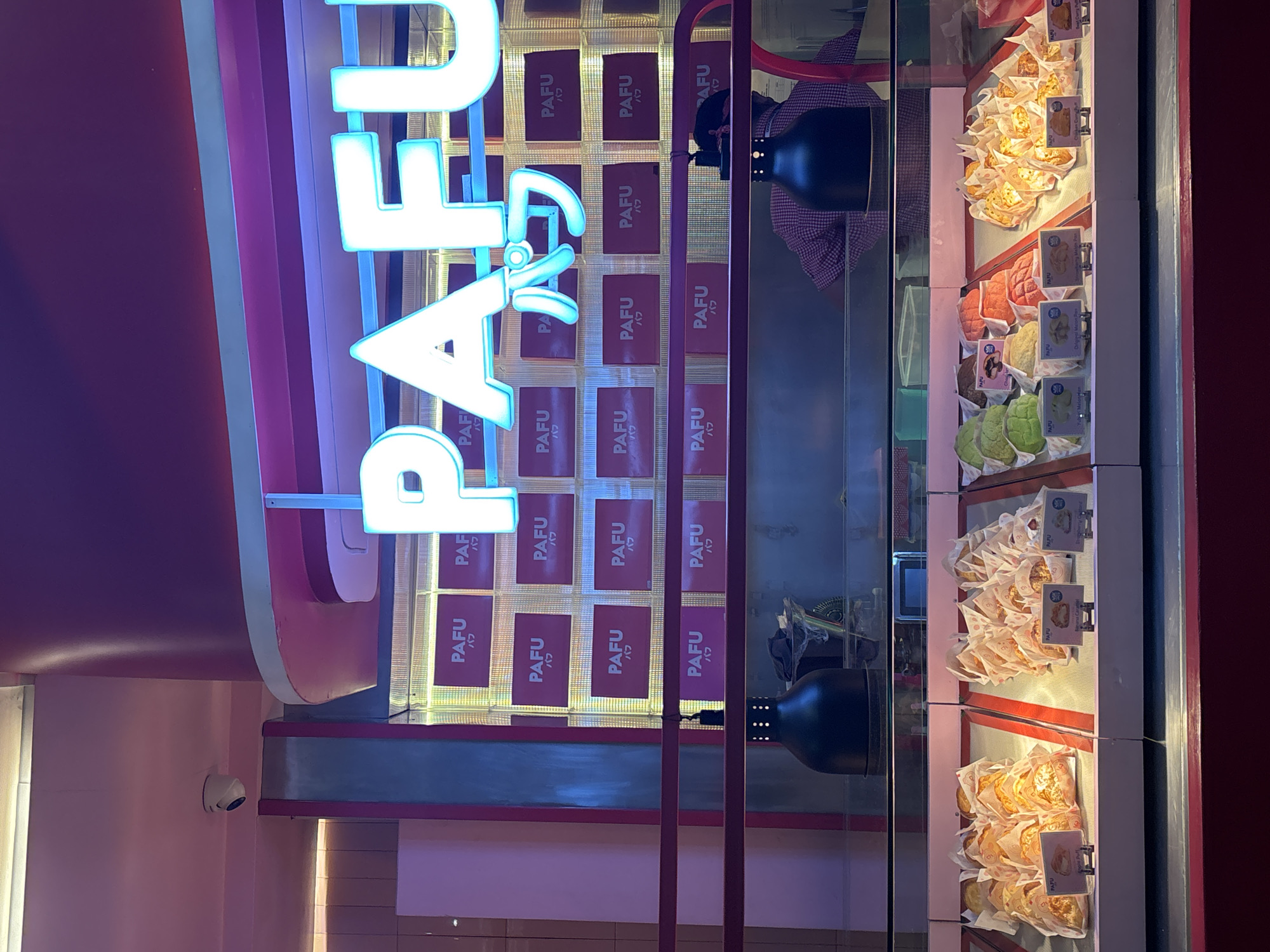 The image shows a bakery or pastry shop, named "PAFU" (with Japanese script underneath), likely in Kuta, Indonesia. The name is displayed in large, illuminated blue letters vertically down the left side of the frame. Behind the letters is a wall covered with repeating logos or labels of the brand "PAFU". A display case filled with various baked goods, including pastries and melon bread, is visible on the right. The bakery appears to have a pink and red color scheme, with a security camera mounted on a wall in the background.