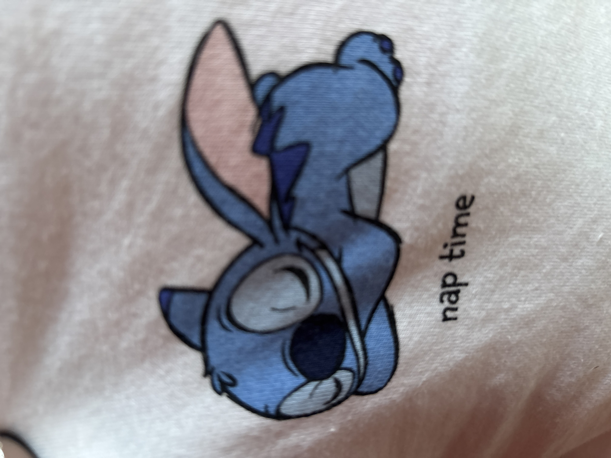 A close-up, slightly angled shot shows a light pink fabric, likely a t-shirt, with a cartoon image of Stitch from Disney's Lilo & Stitch. Stitch is depicted in a relaxed, sleeping pose, lying on his side with his eyes closed and a content expression. He is a vibrant blue with large, rounded ears, one of which is partially visible and has a pink interior. A dark blue bow tie is visible on his back. Below the character, the text "nap time" is printed in black, lowercase letters. The fabric shows a textured weave. The image does not contain any information or visual cues related to Enevo, Bulgaria.