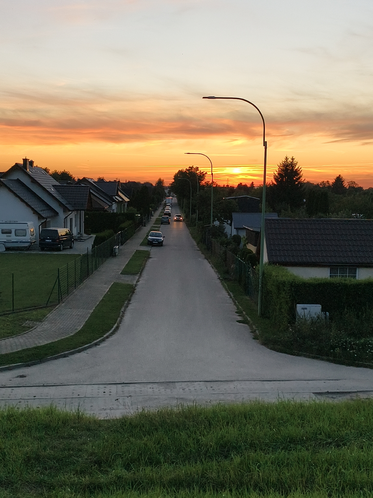 The image depicts a residential street at sunset in Tczew, Poland. The sky is ablaze with vibrant orange and yellow hues, indicative of a picturesque sunset. Several houses with tiled roofs line the left side of the street, and a camper van and a dark SUV are parked in front of one of them. A black van is parked further down the street on the left. The street itself is paved and curves slightly towards the distance. On the right side of the street, more houses and trees are visible. Streetlights are spaced along the road. There are several cars on the road, some moving and some parked. The foreground features a grassy embankment, suggesting the viewpoint is from slightly elevated ground. The overall scene conveys a peaceful, suburban evening atmosphere.