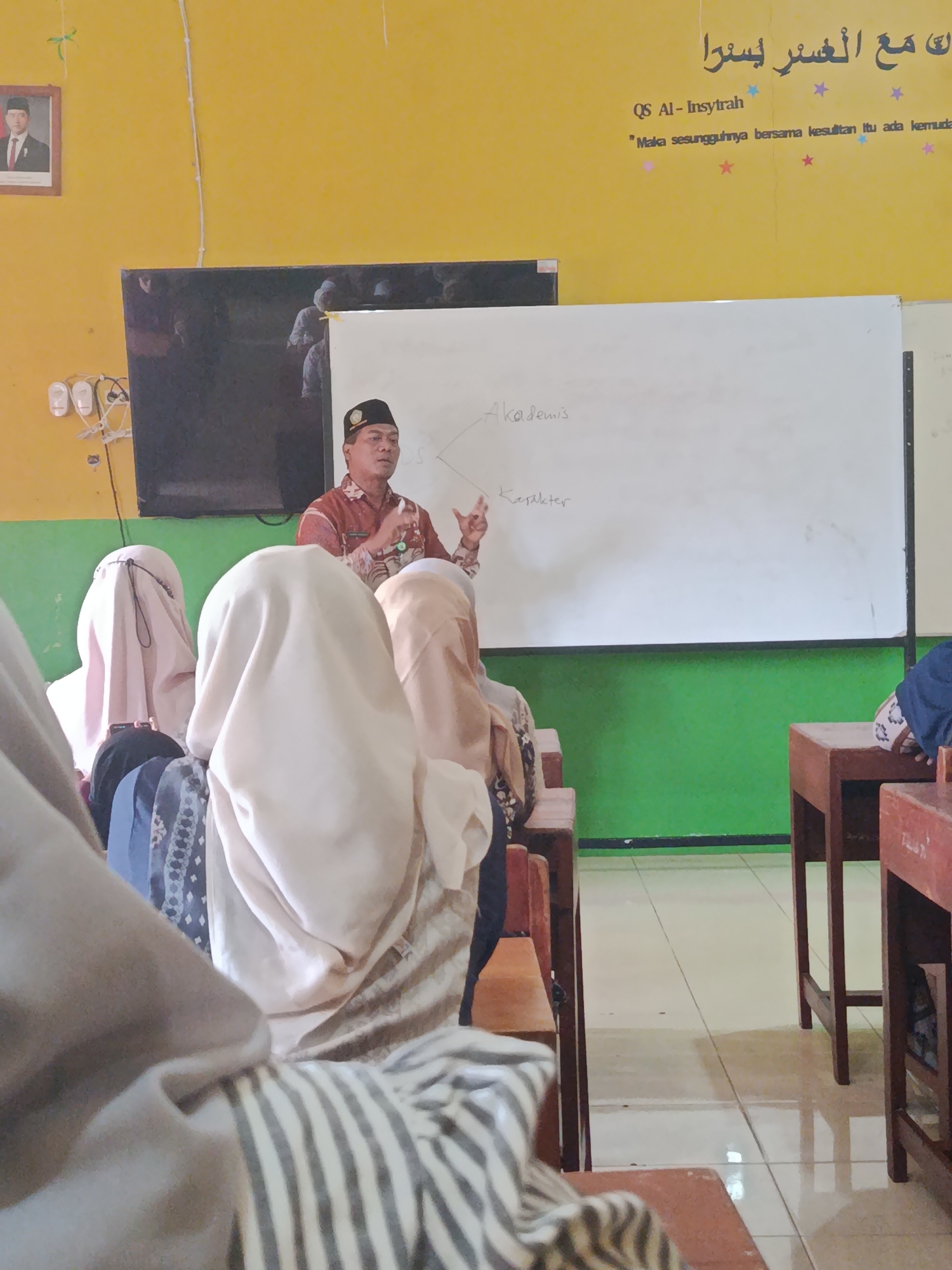 A teacher, wearing a patterned long-sleeved shirt and a black cap, stands in front of a whiteboard in what appears to be a classroom. He is gesturing with both hands while speaking. The whiteboard has handwritten text, including "Akademis" and "Karakter." In the foreground, several students, all wearing hijabs, are seated, their backs to the camera. The classroom has yellow walls, a green section lower down, and a tiled floor. A framed portrait hangs on the yellow wall to the left, depicting a man in formal attire. Arabic calligraphy and a quote in Latin script are also visible on the yellow wall above the whiteboard. The scene is likely located in Kawunganten Lor, Indonesia, given the context provided.