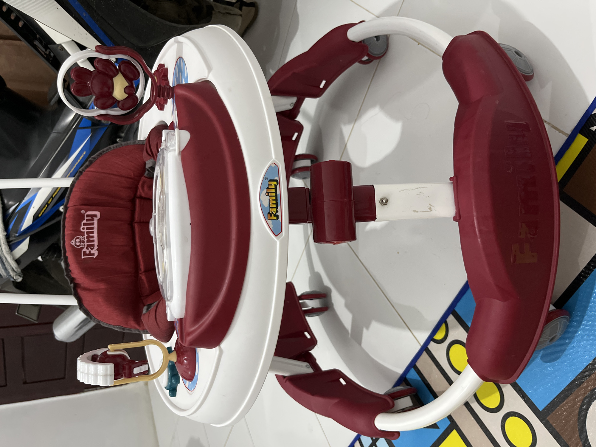 The image shows a baby walker, brand "Family", with a white and maroon plastic frame, a maroon fabric seat, and multi-directional wheels. It's positioned indoors on a tile floor with a colorful, patterned mat partially visible. The location is suggested to be Lhokseumawe, Indonesia, though the indoor setting lacks specific visual cues to confirm the city.