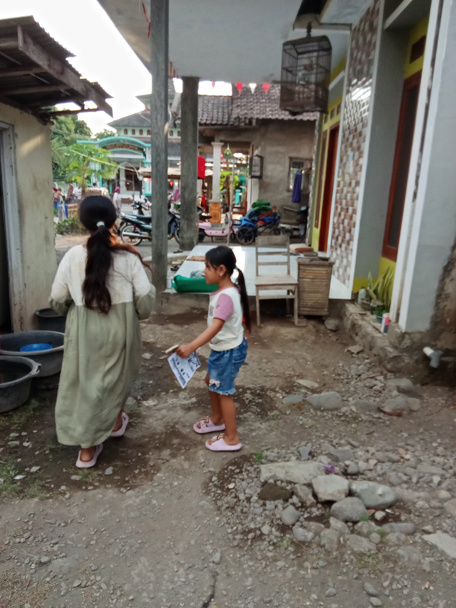 The image depicts an outdoor scene in Selowogo, Indonesia, featuring two girls. The girl on the left, viewed from behind, has dark hair tied in a ponytail and wears a long, light-colored garment. She stands near several large, dark-colored buckets. The girl on the right, facing left, has dark hair in a ponytail and wears a t-shirt with pink sleeves and blue denim shorts. She is holding a folded piece of paper. The environment includes a dirt and gravel ground, with some scattered rocks and a small patch of grass. In the background, there are buildings, one with a patterned facade, and a birdcage hanging from an overhang. Motorcycles are also visible in the background, suggesting a residential or semi-commercial area. The lighting suggests it is late afternoon or early evening.