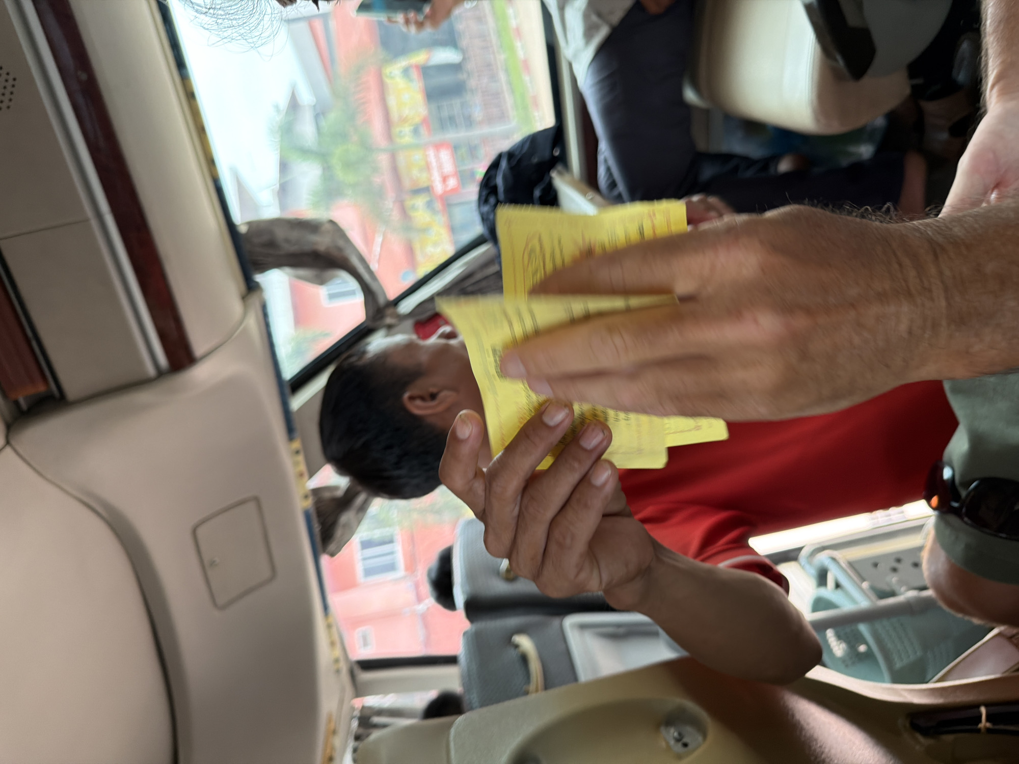 The image shows the interior of a bus, likely in the city of Phakhao, Laos. In the foreground, a person's hands are holding several yellow tickets. The hands are hairy and appear to be Caucasian. Behind the hands, a person wearing a red shirt is visible, holding more yellow tickets. They are sitting on a beige seat with dark grey upholstery. Other passengers are visible in the background, as are the windows of the bus, which offer a view of buildings and palm trees outside. The overall scene suggests a travel context.