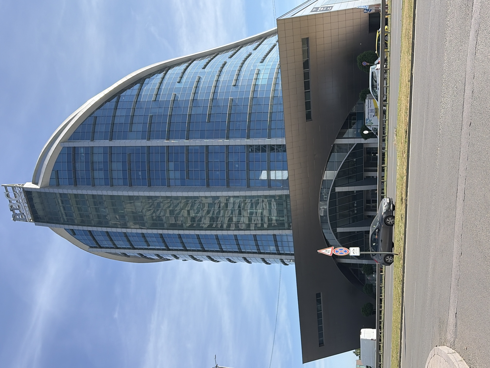 In Sofia, Bulgaria, a modern, curved glass skyscraper with the "BBC" logo at its apex dominates the skyline. The building features a sleek, metallic lower section that extends outwards over an arched entrance. In front of the building, a multi-lane road runs horizontally, with a grassy median separating traffic. Several vehicles, including a white van and two cars, are visible on the road and the adjacent shoulder. A traffic sign with "no stopping" and "no parking" symbols stands near the roadside. The sky is clear and blue with scattered wispy clouds, indicating a bright, sunny day.
