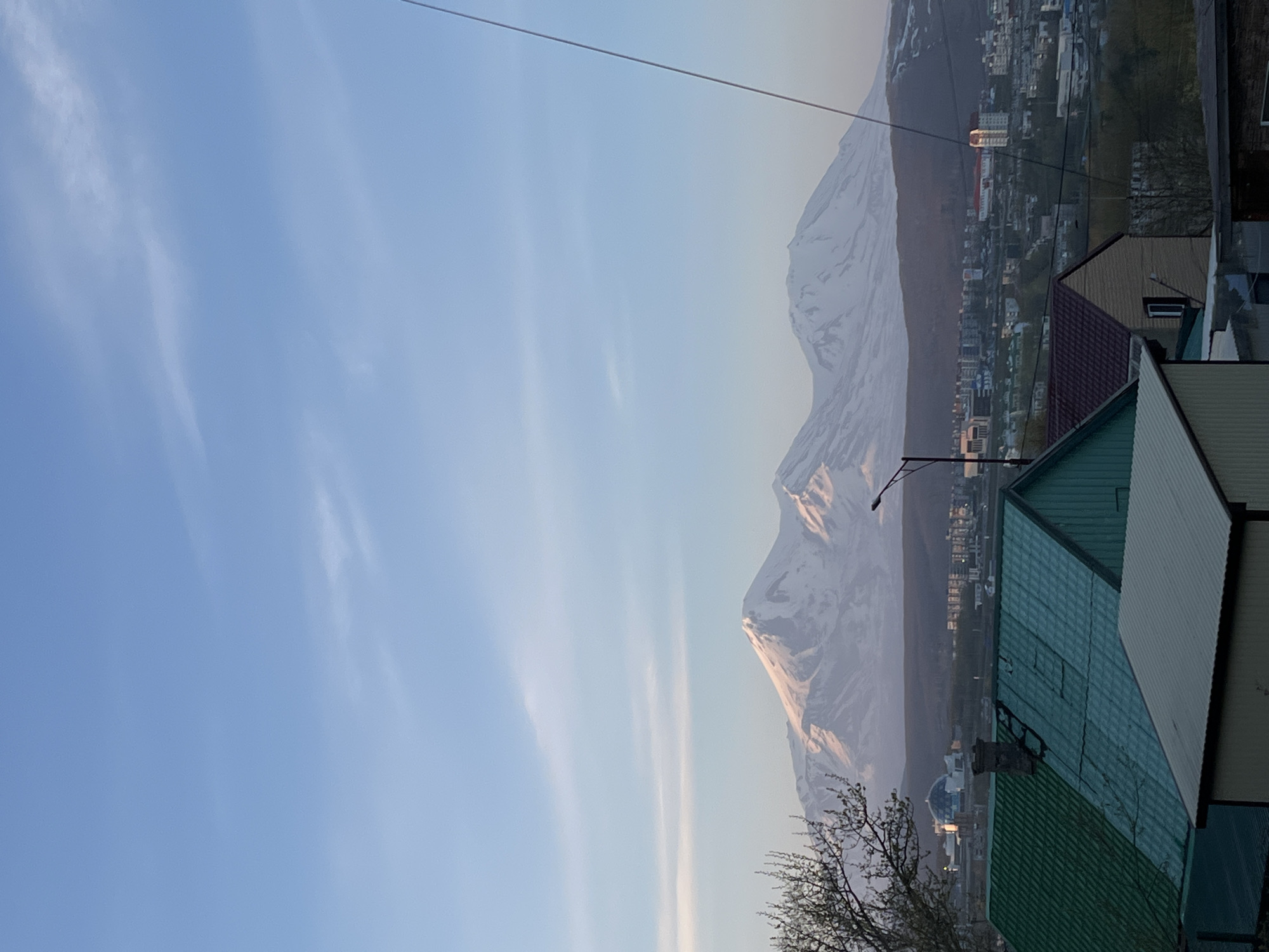 This landscape image captures a panoramic view of Petropavlovsk-Kamchatsky, Russia, with its distinct mountainous backdrop, likely taken during early morning or late afternoon.
In the foreground, the image features the roofs of several buildings, mostly residential or utilitarian structures, with varying colors like teal, dark green, and brown. Bare tree branches are visible on the left, suggesting a season other than full summer. A thin power line stretches across the upper part of the frame.
The midground reveals a sprawling urban area with numerous buildings of different sizes and styles. A prominent circular, dome-shaped building is noticeable towards the left side of the city, and a streetlamp is also visible.
Dominating the background are majestic, snow-capped mountains. The snow on their peaks is illuminated by a warm, golden light, indicating either sunrise or sunset. The mountains appear rugged and volcanic, characteristic of the Kamchatka region.
The sky above is a clear, light blue with scattered, wispy white clouds, further contributing to the calm and serene atmosphere. No people or specific activities are visible in the frame, and there is no discernible text. The scene conveys a sense of grandeur and the striking coexistence of urban development with dramatic natural landscapes.