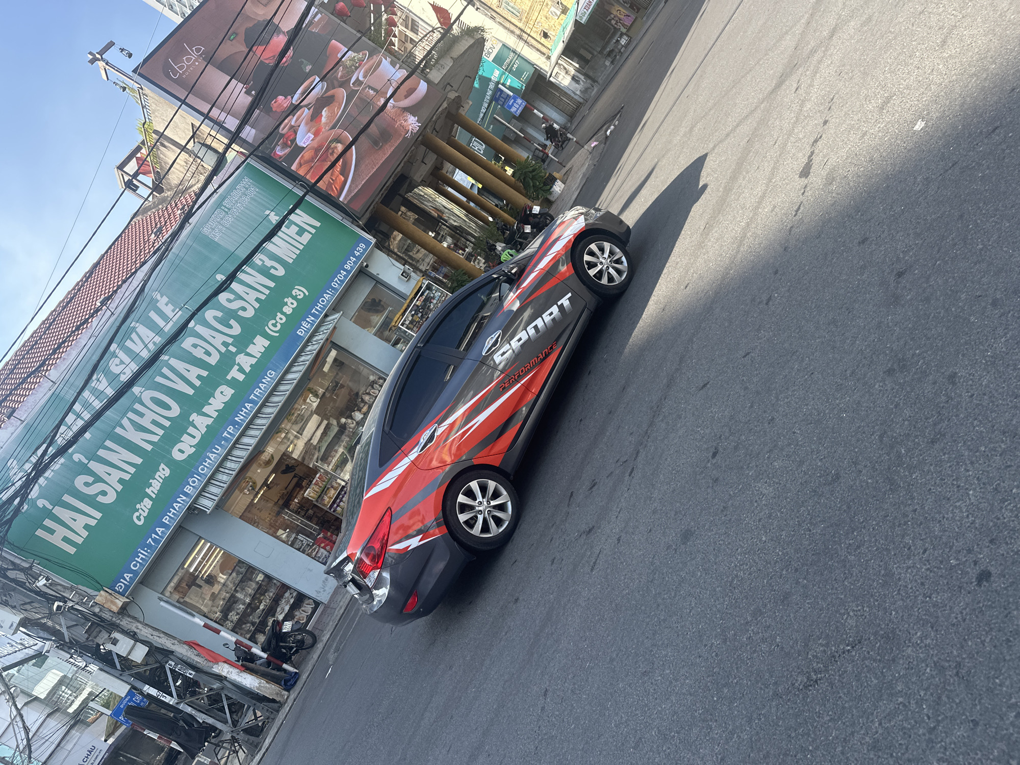 In Nha Trang Ward, Vietnam, a dark gray car with red and white racing stripes and the word "SPORT" emblazoned on its side is parked on a street. The car is positioned at an angle, with its rear end closer to the foreground. Behind the car, a green banner with white Vietnamese text hangs above a storefront, advertising "HẢI SẢN KHO VÀ ĐẶC SẢN 3 MIỀN" (Dried Seafood and Specialties from 3 Regions) and providing an address: "ĐỊA CHỈ: 71A PHAN BỘI CHÂU - TP. NHA TRANG" (Address: 71A Phan Boi Chau - Nha Trang City) and a phone number: "ĐIỆN THOẠI: 0704 904 439" (Phone: 0704 904 439). Adjacent to this banner is a large advertisement for "ibata HOTELS&SPA" featuring images of food. The street is paved and appears to be a busy commercial area, with utility poles and wires overhead, and other businesses visible in the background. The sky is clear and blue.